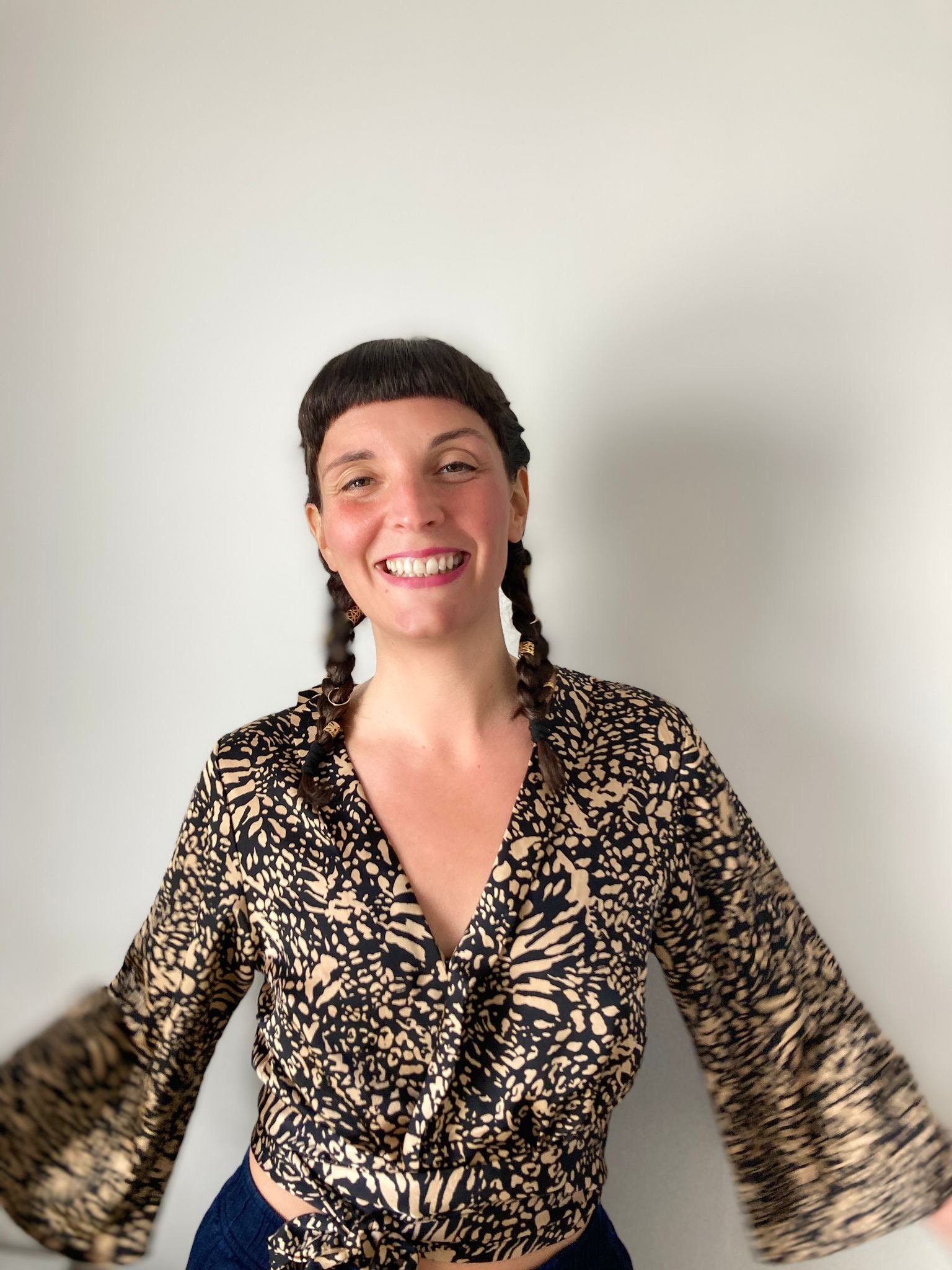 Young woman with dark hair in braided pigtails, smiling, wearing a leopard print blouse, standing against a plain white wall.