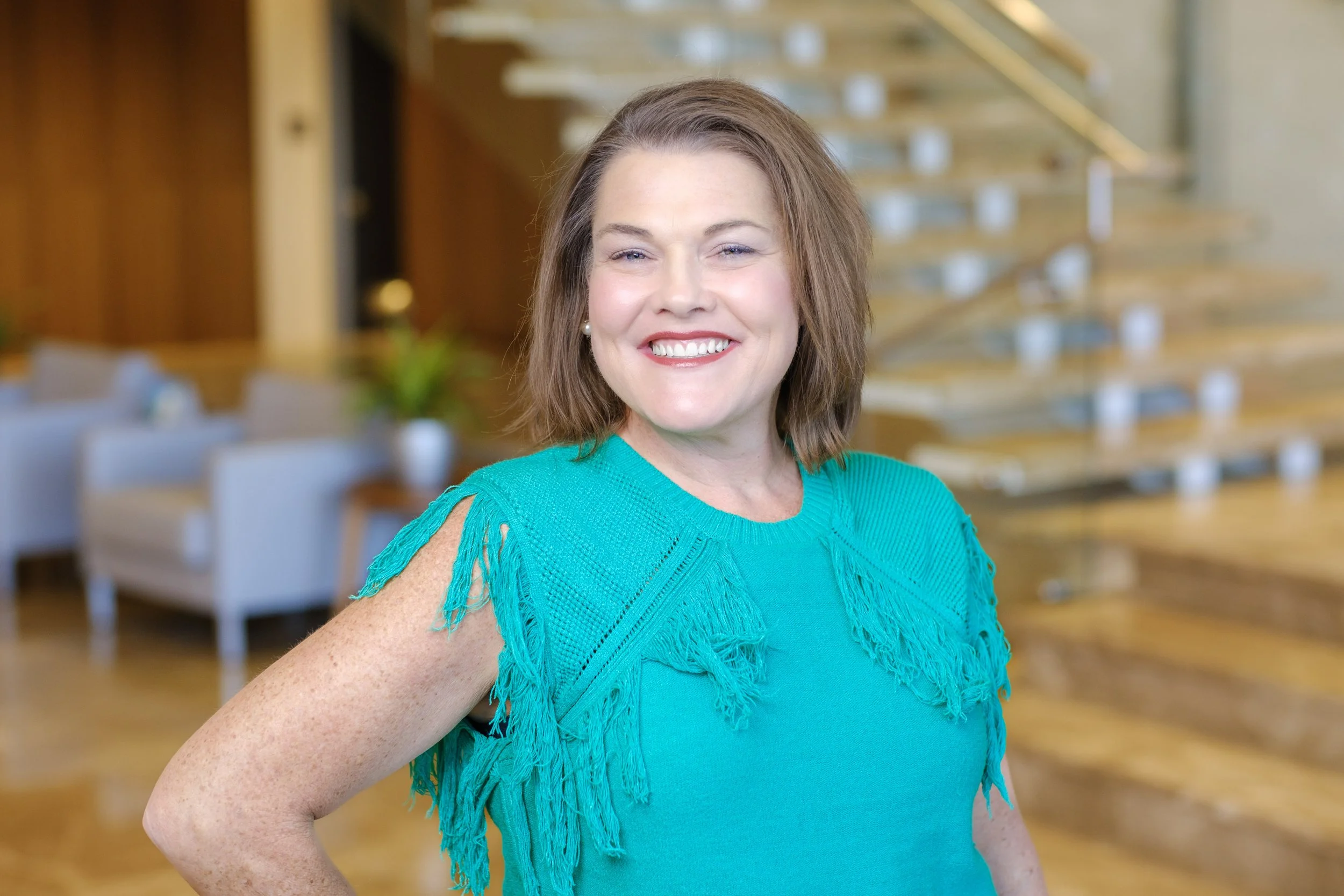 A woman with brown hair, wearing a teal blouse with cut-out shoulders, smiling in a brightly lit, modern indoor space with wooden stairs and white chairs in the background.