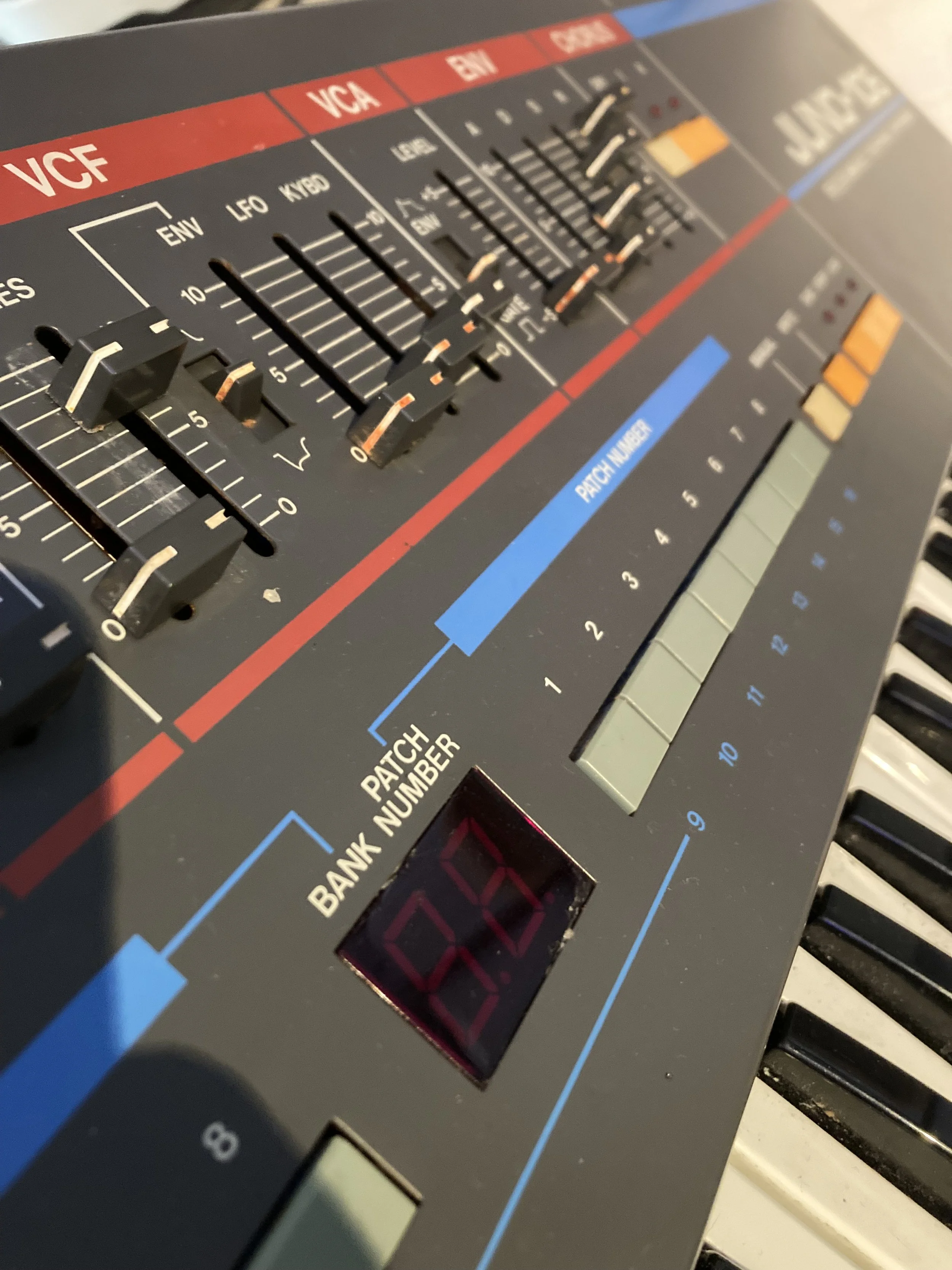 Close-up of a vintage Juno 106 keyboard synthesizer showing knobs, sliders, and buttons with labels for sound adjustments such as VCF, VCA, ENV, LFO, KBD, and PATCH NUMBER, with part of piano keys visible at the bottom.