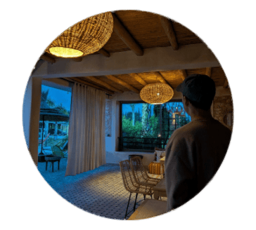 A person looking out a large window in a cozy, well-decorated room with wicker furniture and hanging woven lamps.