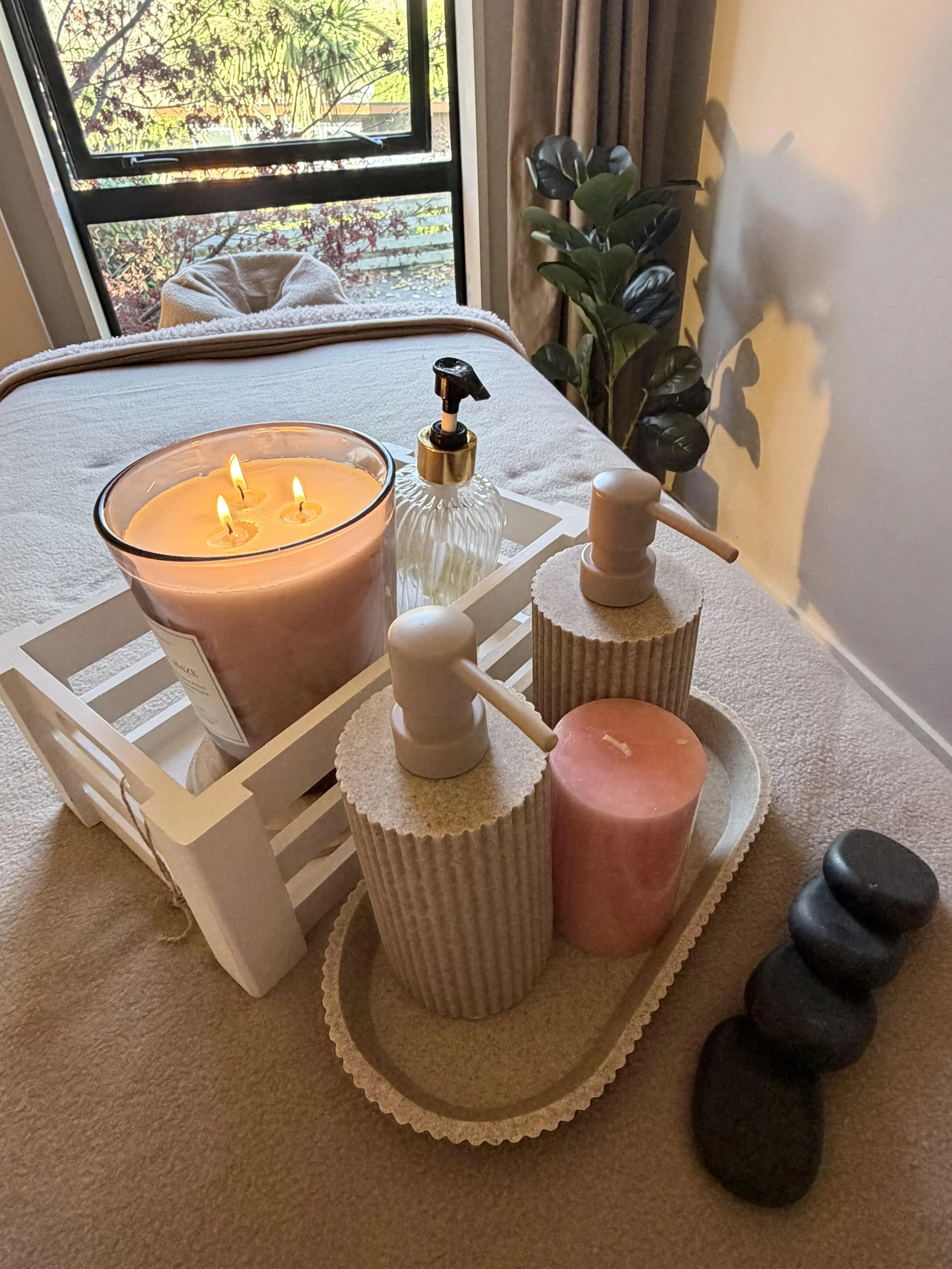 A cozy setting on a table with candles, a soap dispenser, and decorative objects near a window with a view of trees and plants outside.