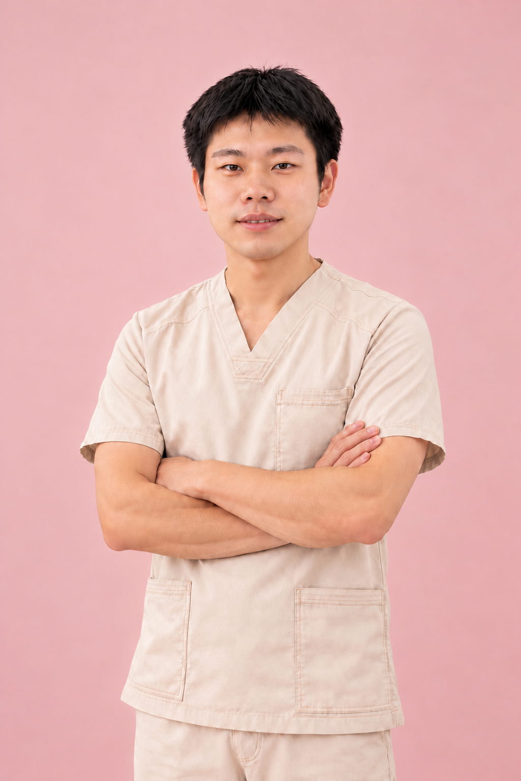 A young man with short black hair wearing beige medical scrubs with arms crossed, standing against a pink background.