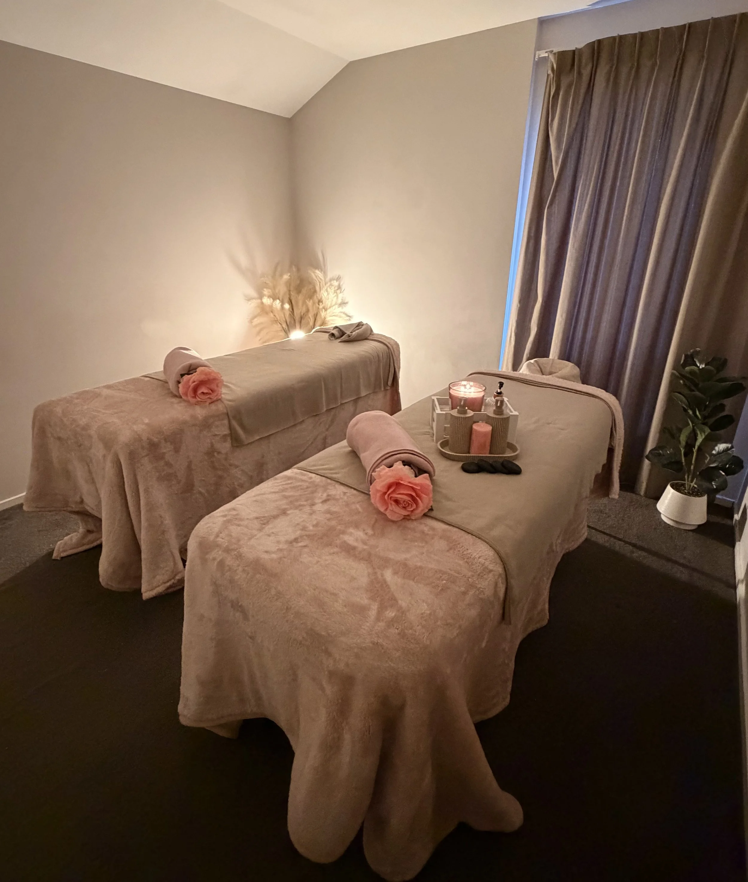 A massage table covered with a beige sheet, with pink roses, candles, and massage oils on a tray. There is a window with closed brown curtains, a potted plant, and a decorative feather lamp.