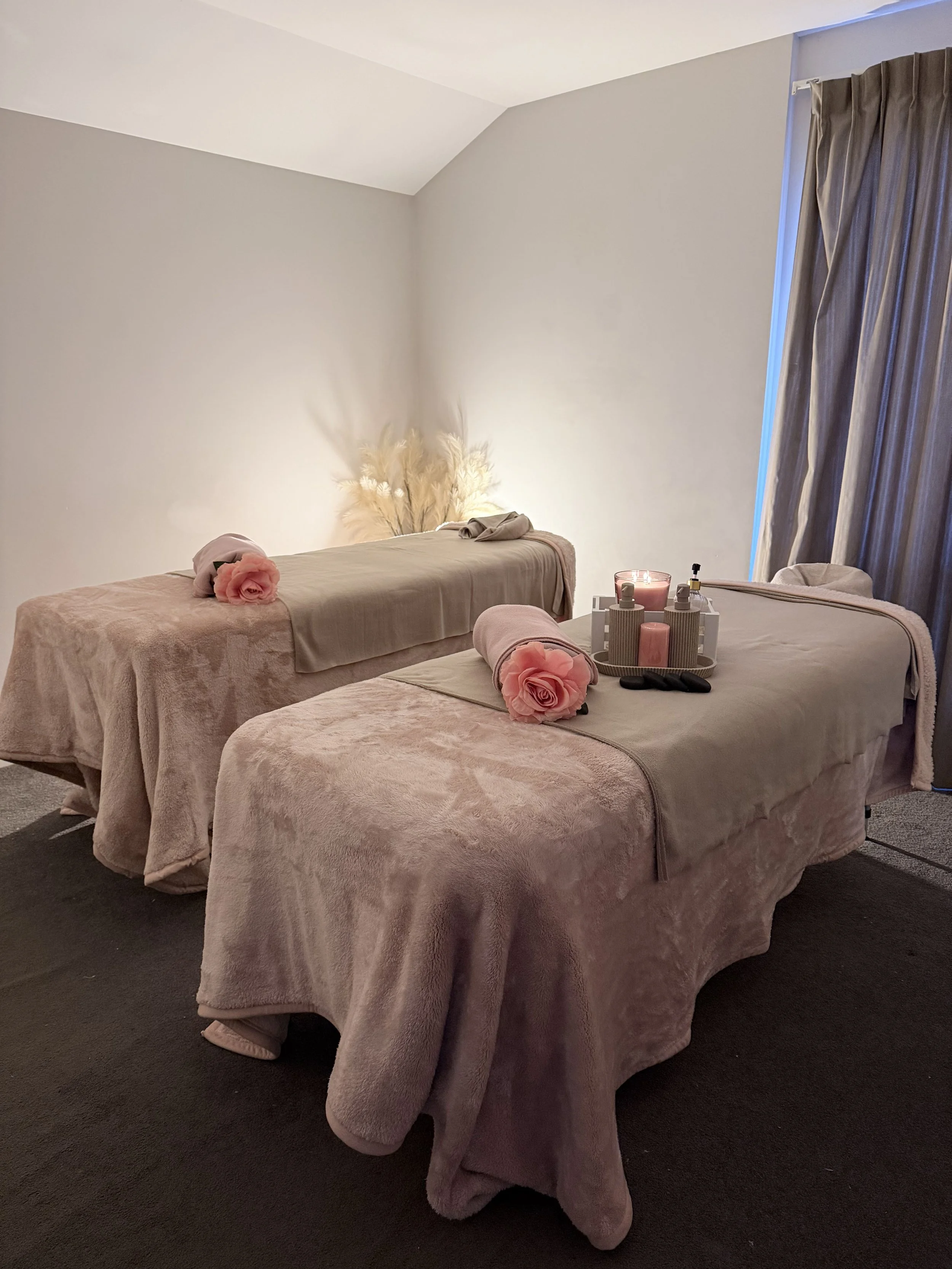 A room with two massage tables covered in pink plush blankets and rolled towels with pink roses on top, set against a plain white wall, with a window hidden behind gray curtains, and decorative pampas grass in the corner.