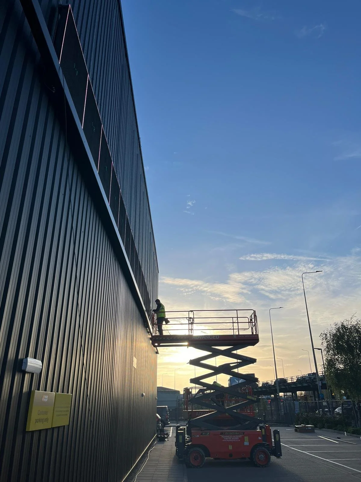 A worker is on a scissor lift performing maintenance or installation work on a large, dark, corrugated metal building. The sky is partly cloudy with the sun low in the sky, indicating early morning or late afternoon.