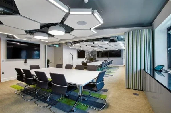 Modern conference room with a large white table surrounded by black chairs, wall-mounted flat-screen TVs, geometric ceiling lighting, a coat rack, and a window with curtains.