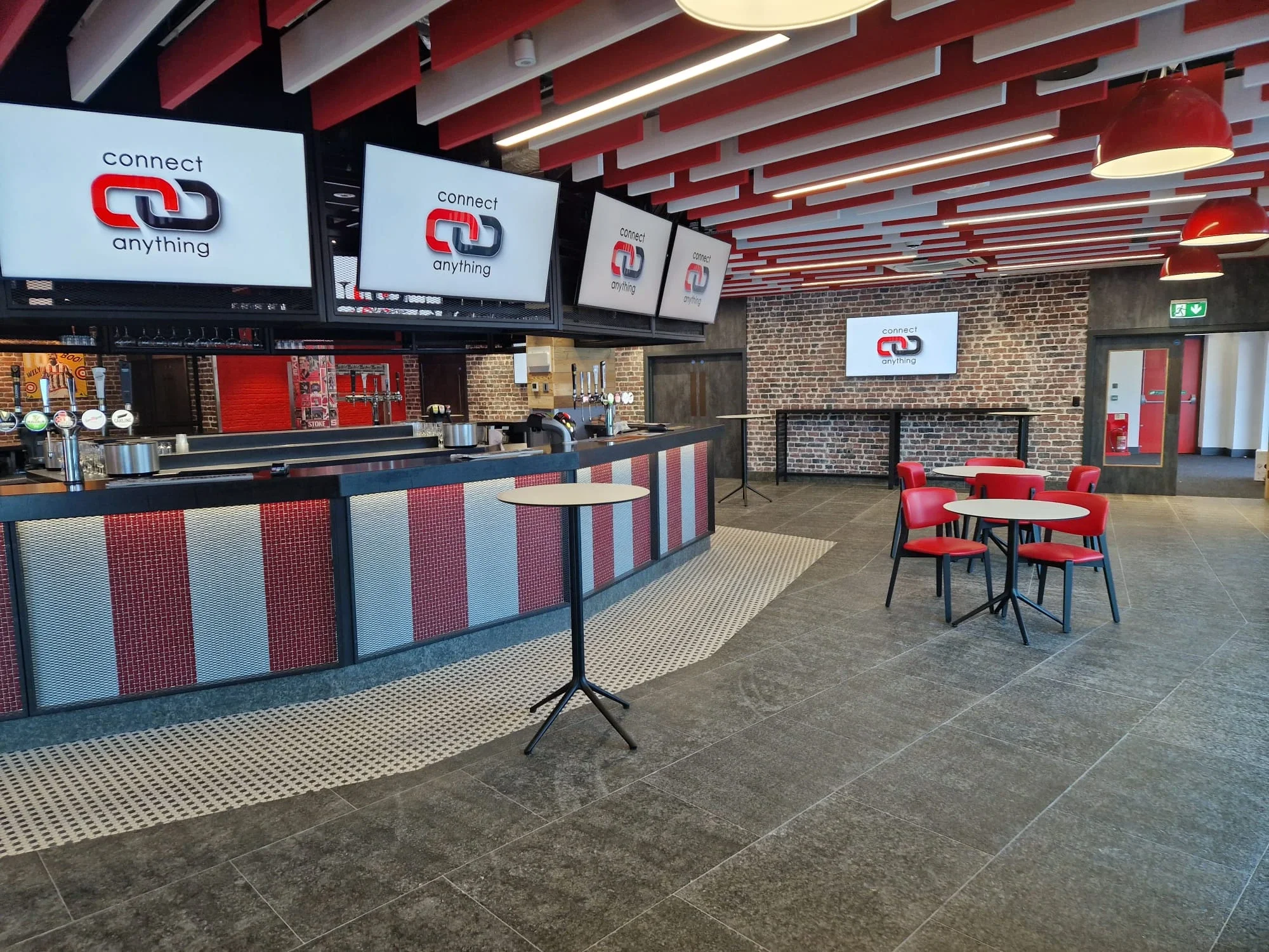 Modern bar with a red and black color theme, multiple digital screens displaying the words 'connect anything' with a logo of two interlinked chains, brick wall background, and red chairs around small round tables.