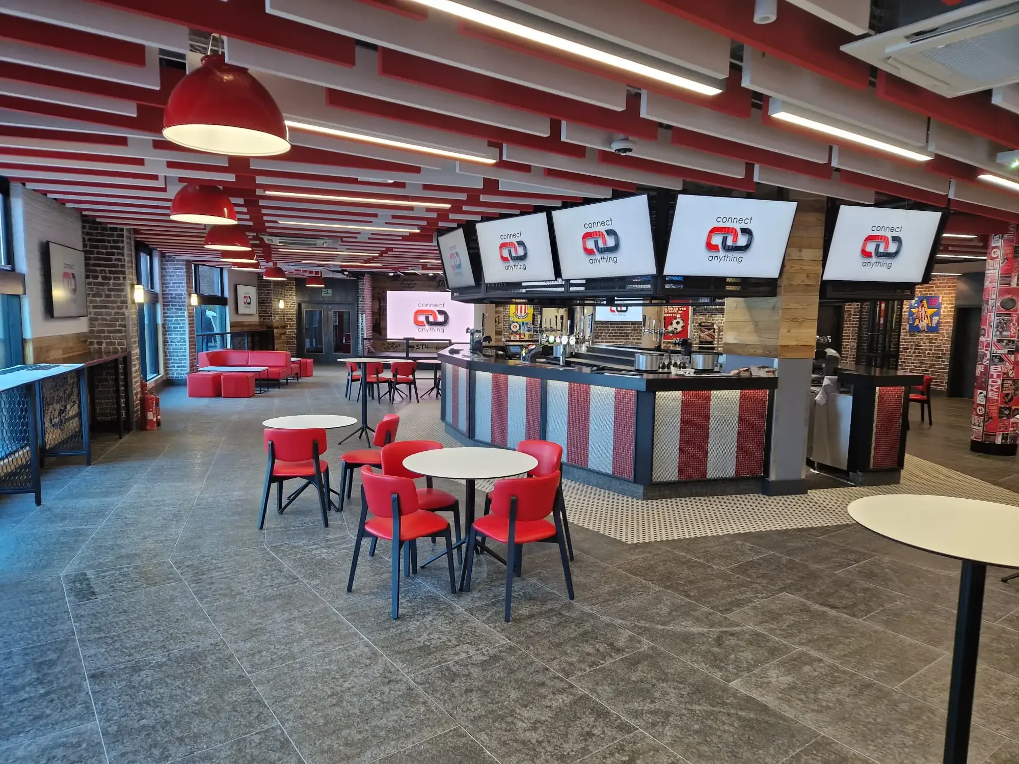 Interior of a modern sports bar or pub with red and black color theme. Red pendant lights, digital screens displaying 'connect anything', red chairs, small round tables, exposed brick walls, and a bar counter with beer taps.