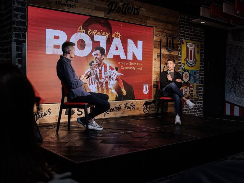 Two men sitting on a stage in front of a large poster advertising an evening with Botan in aid of Stoke City Community Trust. One man is holding a microphone and the other appears to be taking notes or reading. The stage has wooden flooring and is located in a venue decorated with sports-related artwork and branding.