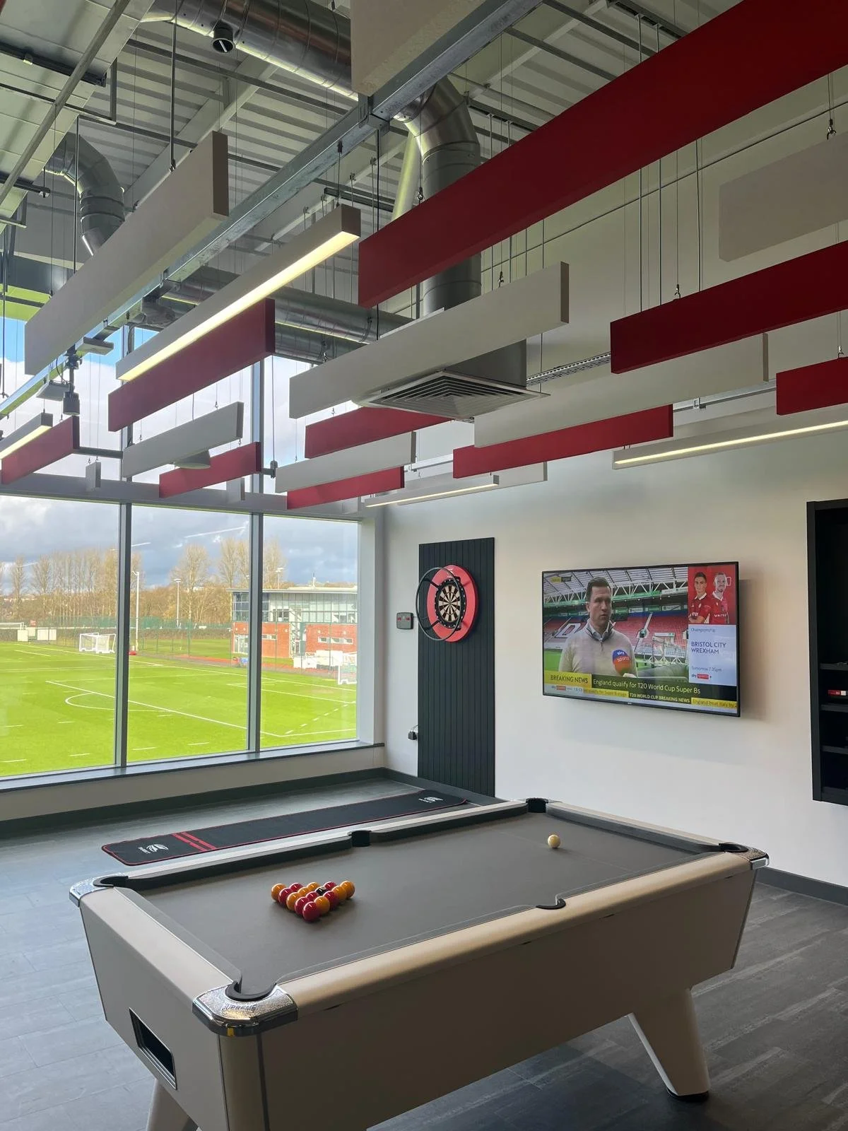 Indoor room with a pool table featuring a triangle of billiard balls and a cue ball. A dartboard is mounted on the wall, and a flat-screen TV shows a sports news broadcast. Large windows reveal a soccer field outside with goals and a building in the background.