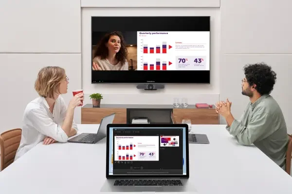 A meeting room with three people, two women and one man, sitting at a white table with laptops, watching a presentation on a wall-mounted screen displaying charts and graphs.