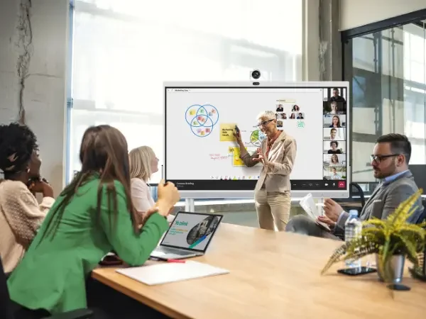 A woman leading a presentation in a conference room, with four attendees listening and taking notes, a large screen displaying diagrams and video thumbnails, and a glass wall letting in natural light.