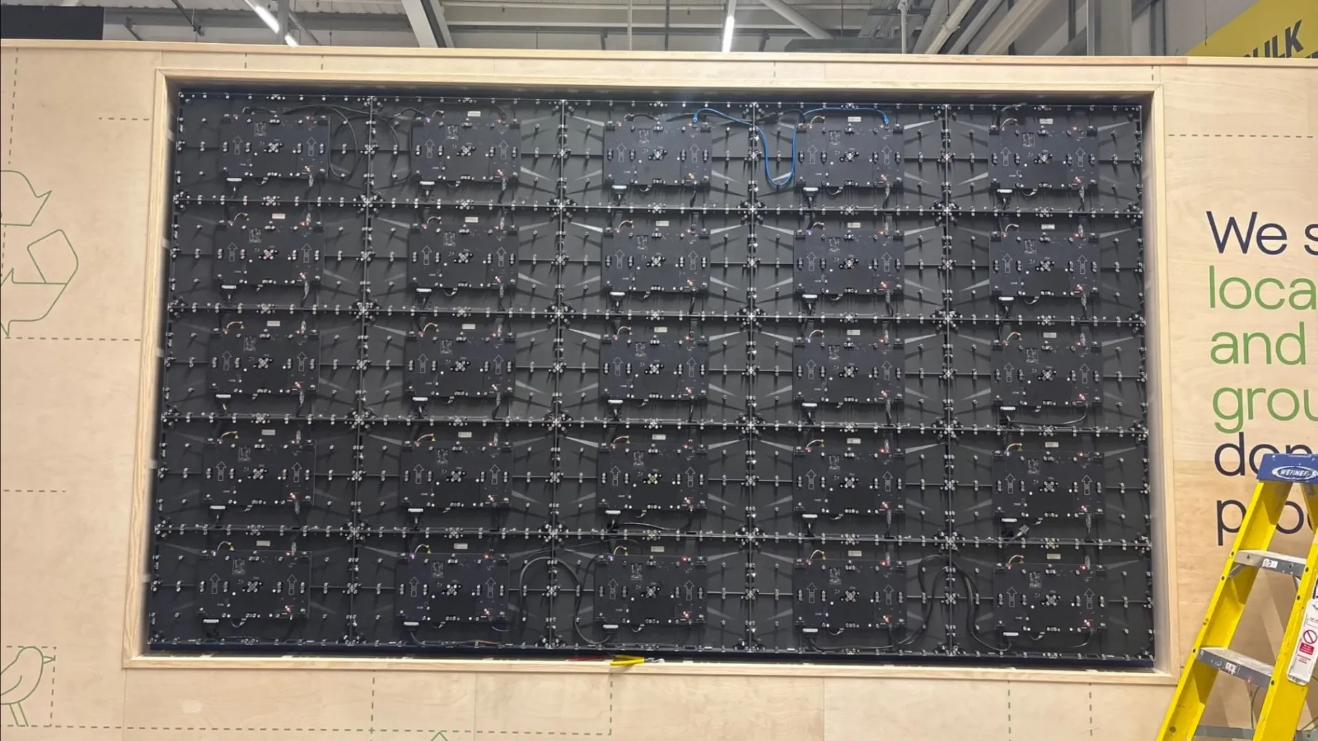 Large wooden frame with an intricate array of black circuit boards and wires mounted on it, with a yellow ladder on the right side.
