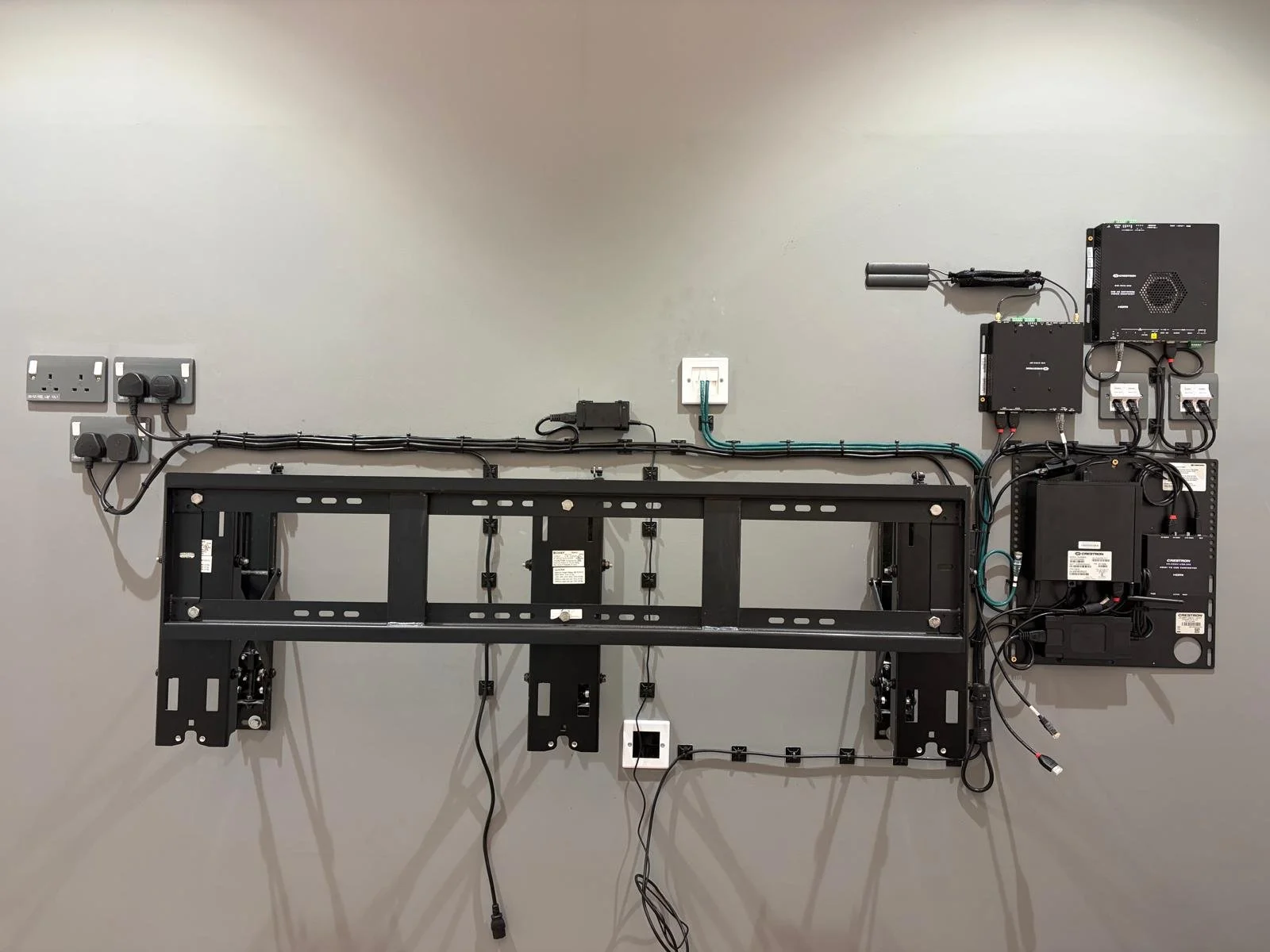 An empty wall mount with connected electrical and electronic components, including power outlets, black modular devices, and wiring, with shadows cast on the wall.
