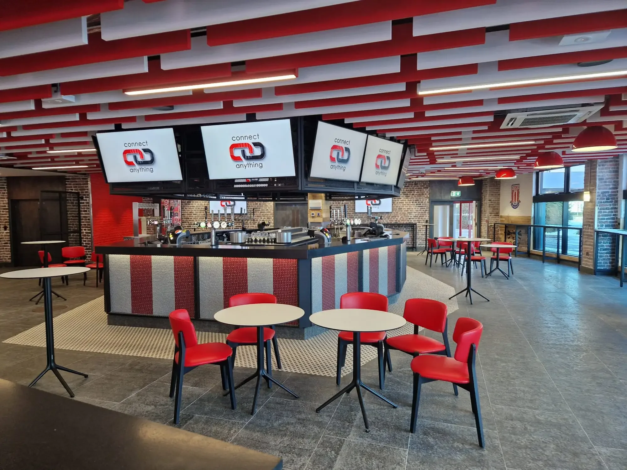 Interior of a modern bar or cafe with red chairs and tables, brick walls, multiple television screens displaying a logo with interconnected red and black shapes and the words "connect anything." The ceiling features red and white wooden slats with in