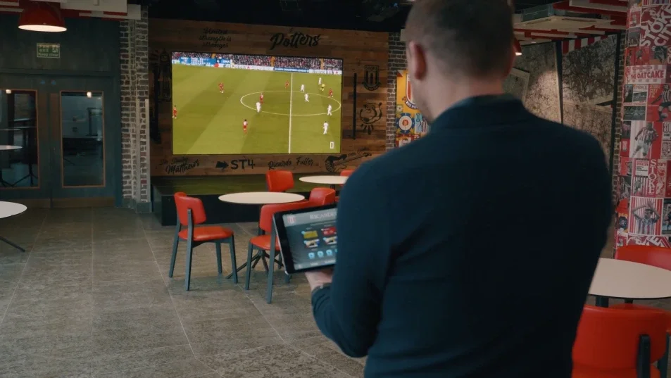 A man in a black shirt watching a soccer game on a large screen TV in a sports bar, holding a tablet.