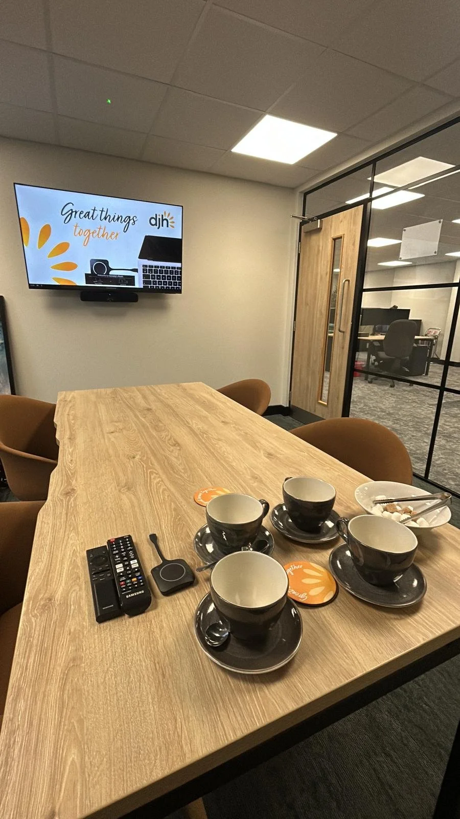 A conference room with a wooden table, four black and white coffee cups with matching saucers, a bowl with a spoon, a TV screen displaying the text 'Great things together' and the logo 'djh', and a door leading to an office area with gray chairs and tables.
