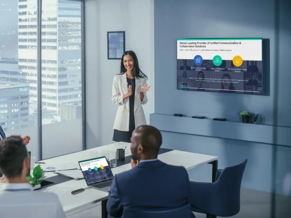A woman giving a presentation in a modern conference room with large windows, while three people in business attire are seated at the table, looking at her and the screen.