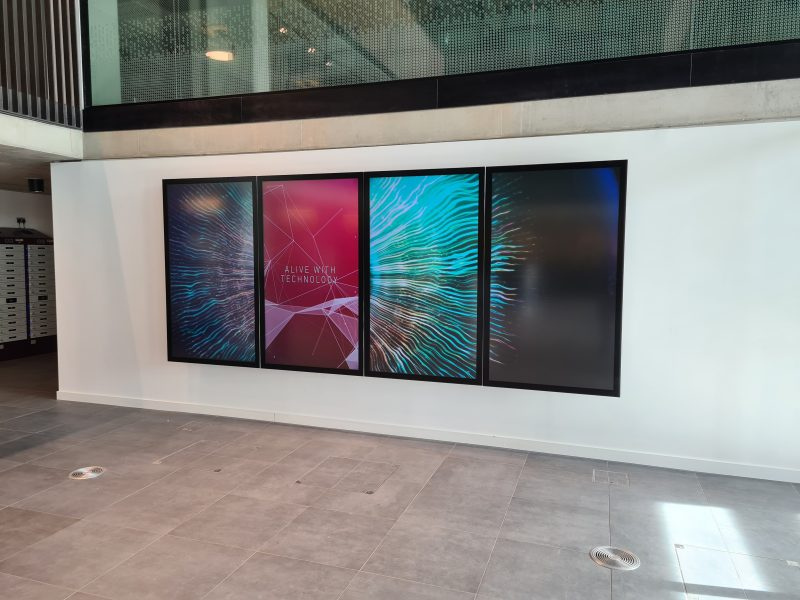 Four digital display screens mounted on a white wall in a modern indoor space. The first three screens show colorful abstract graphics, and the second screen has the text 'ALIVE WITH TECHNOLOGY'.