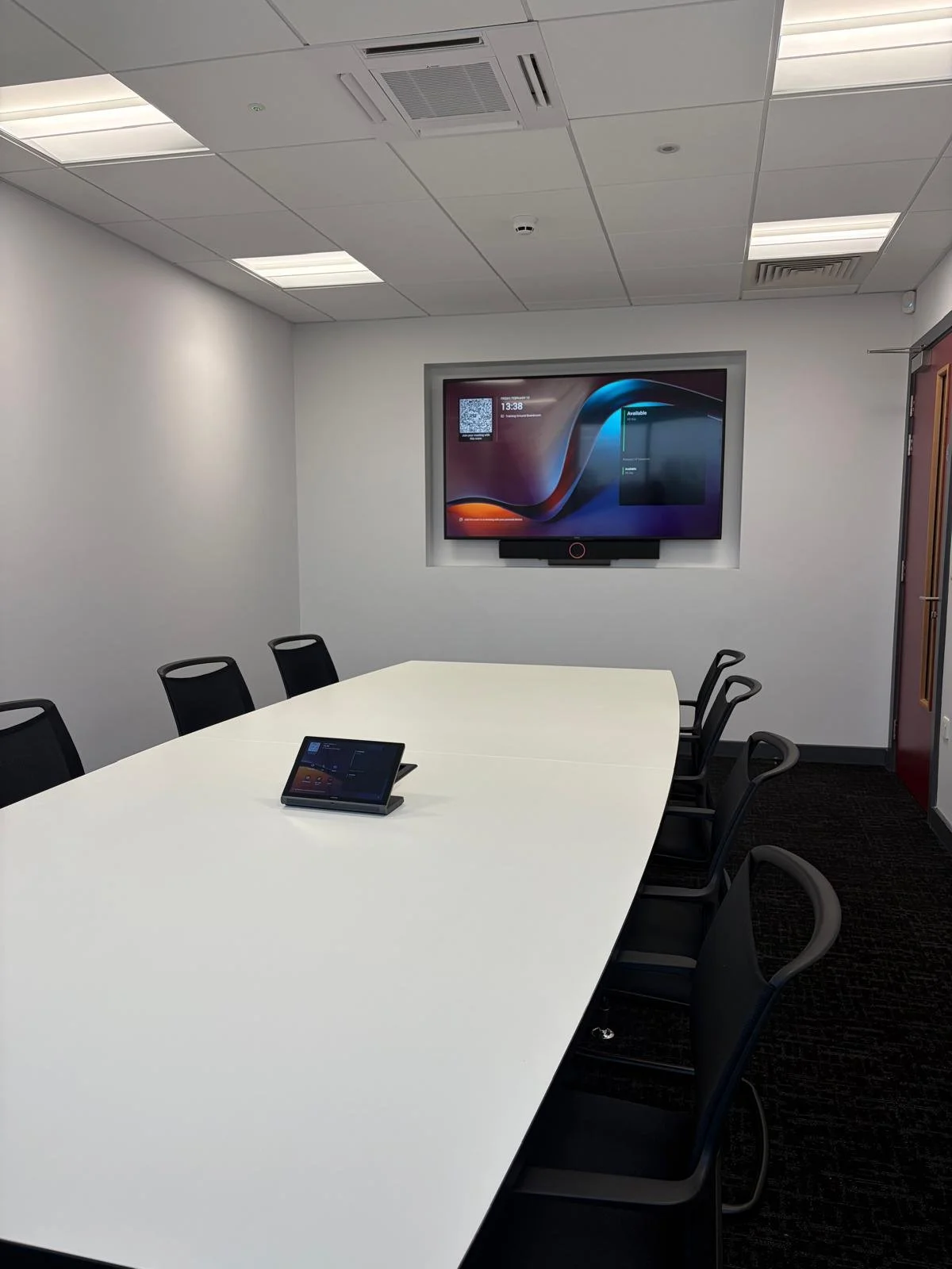 Empty conference room with white oval table, black chairs, a tablet on the table, and a wall-mounted monitor.