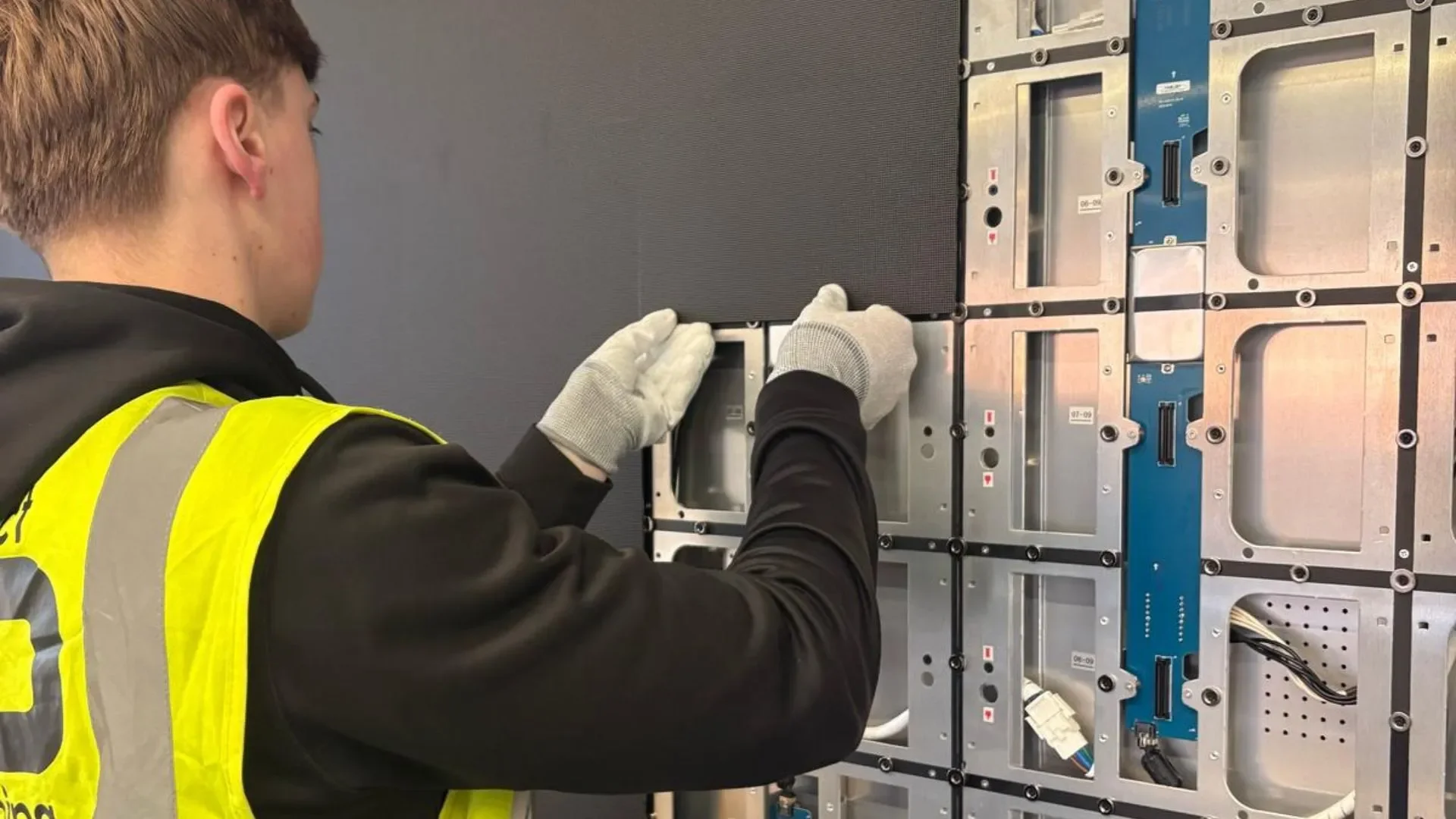 A person wearing a yellow safety vest, white gloves, and a black long sleeve shirt is working on the interior of a large electronic or mechanical device, possibly an industrial machine or computer hardware, mounted on a wall.