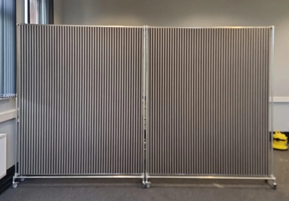 A portable metal room divider with vertical slats, set up indoors with a gray wall and floor.