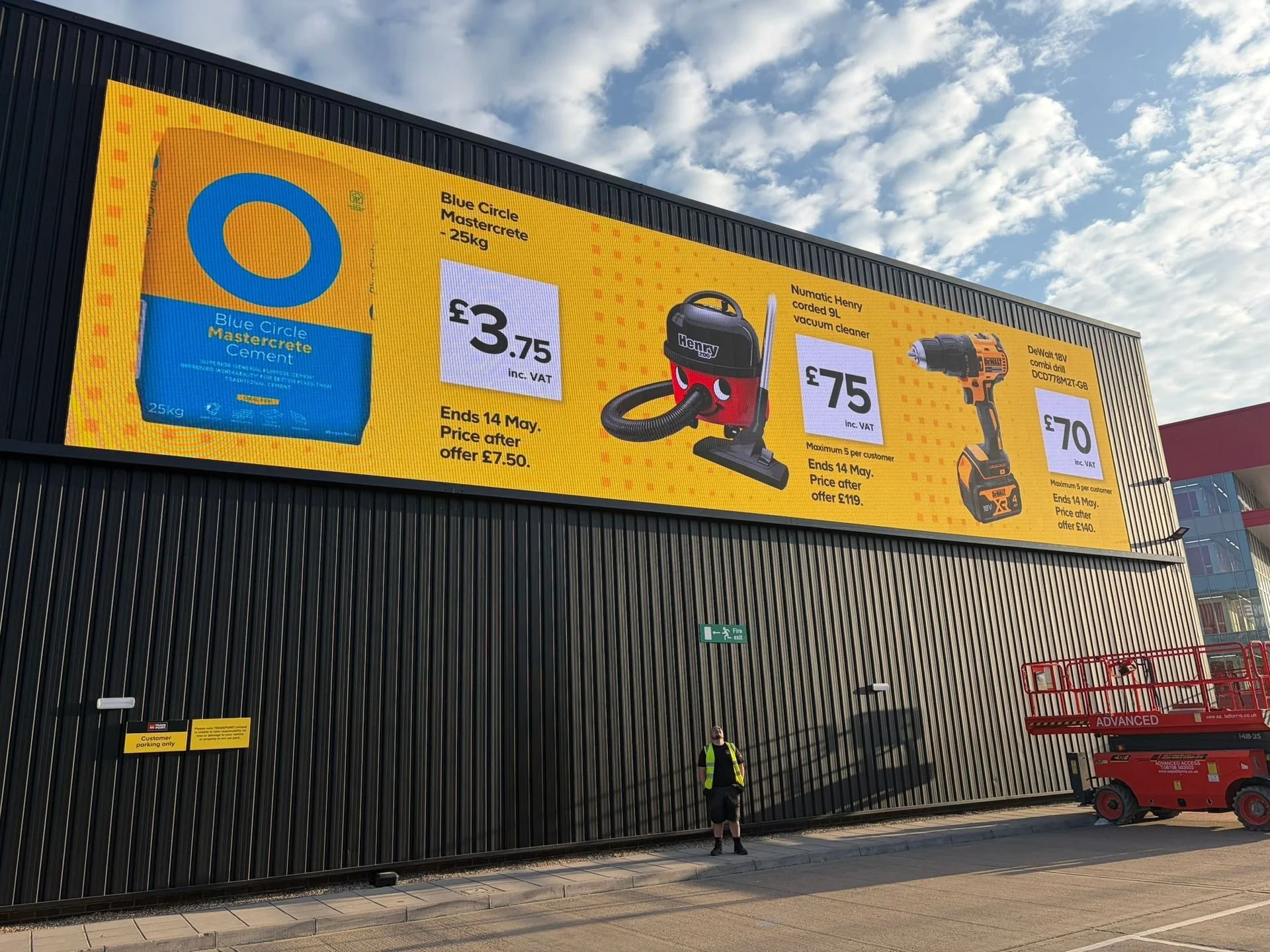 Large yellow advertisement billboard on the exterior of a building, promoting a cement bag, vacuum cleaner, and cordless drill with prices and offers. A man with a high visibility vest and shorts stands below, with a shopping cart and a fire exit sign nearby.
