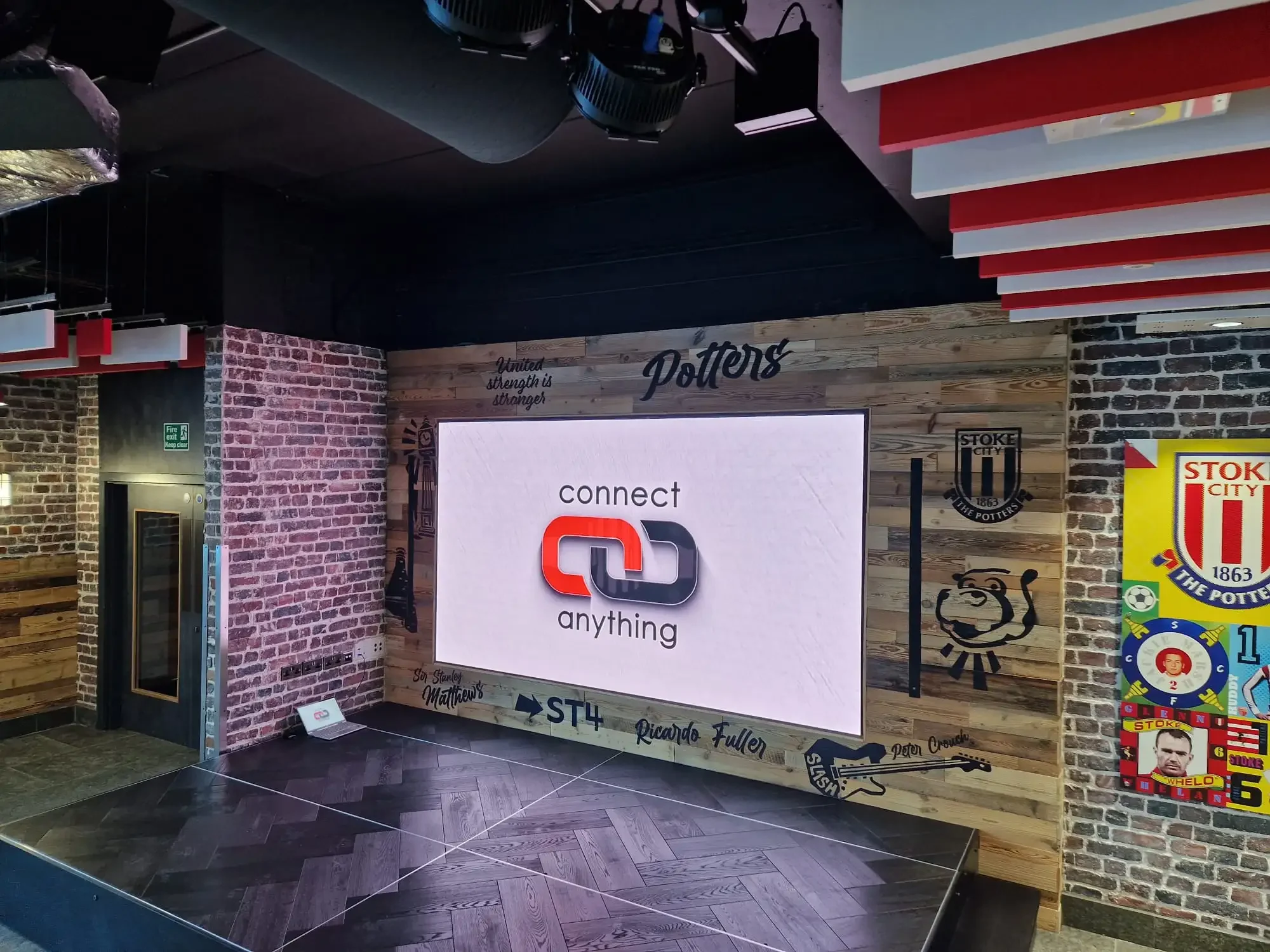 Indoor display area with a large screen that reads 'connect anything.' The wall features a brick design on the left and a wooden panel with various writings and logos, including 'Potters.' There are black and red accents on the ceiling, and a fire ex