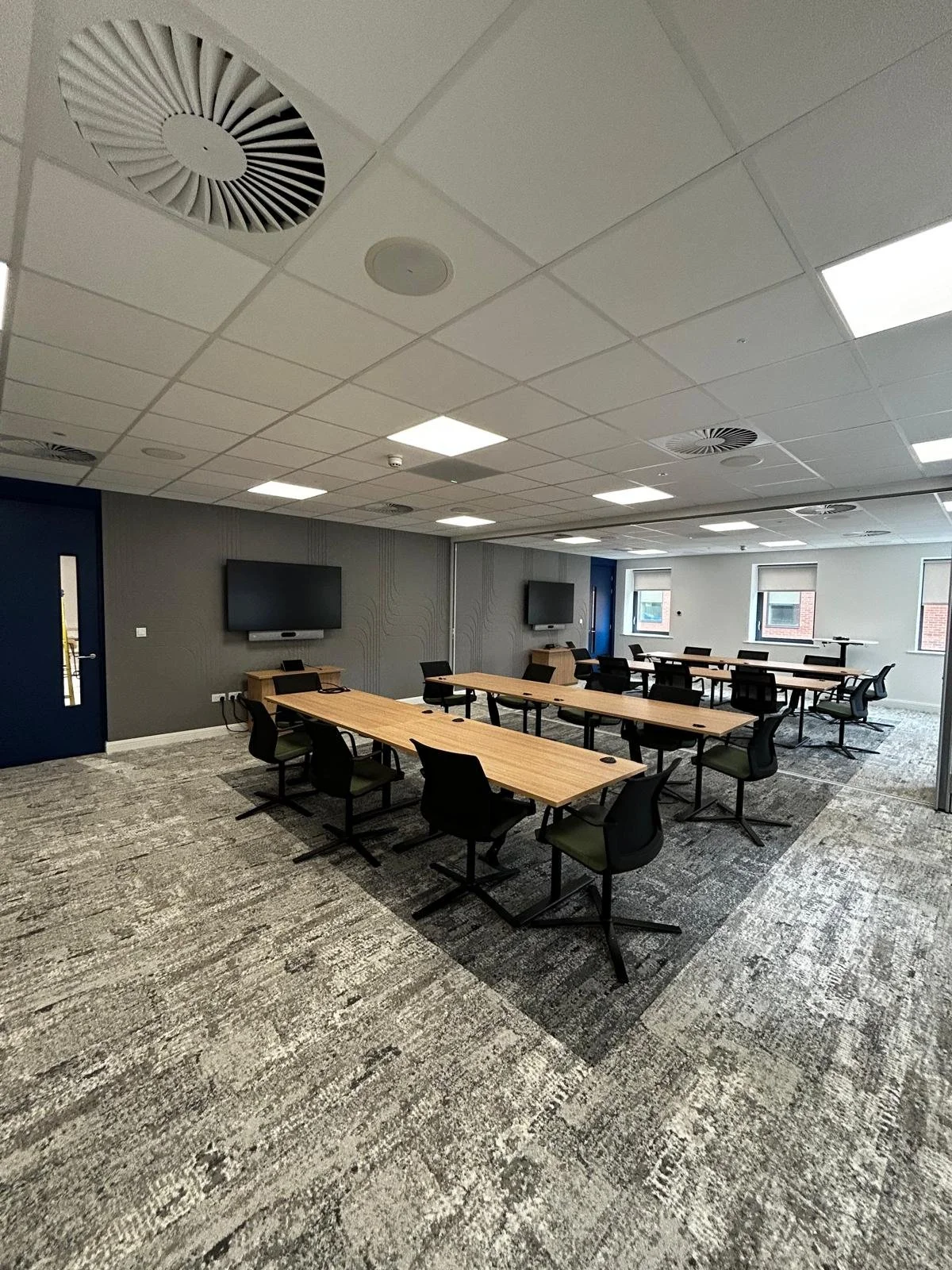 Empty modern conference room with wooden tables, black chairs, wall-mounted screens, and large windows.
