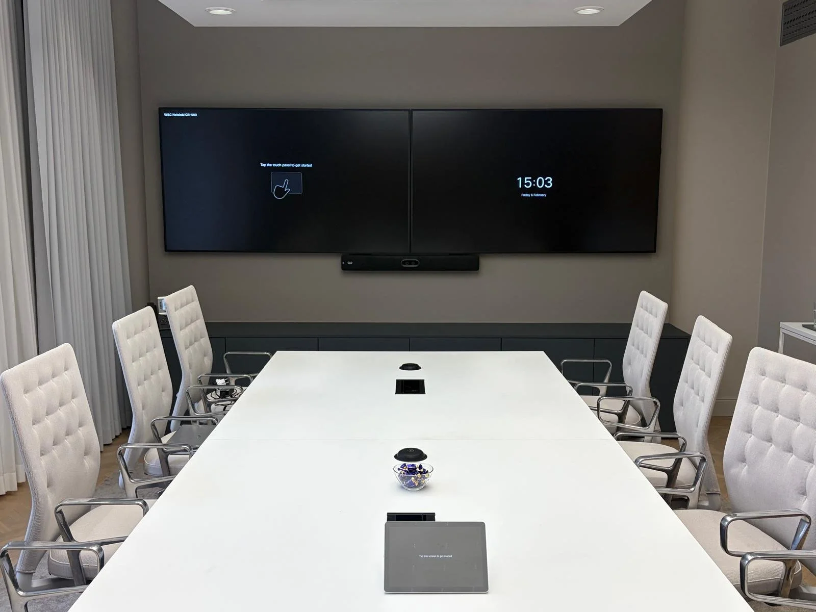 A modern conference room with a long white table, white cushioned chairs with metal armrests, a large wall-mounted screen showing instructions to tap the touch panel to get started and the time 15:03, and a small bowl of candy on the table.