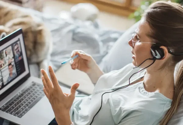 Woman with glasses and headphones using a laptop for a video call, holding a pen, with a cat in the background.