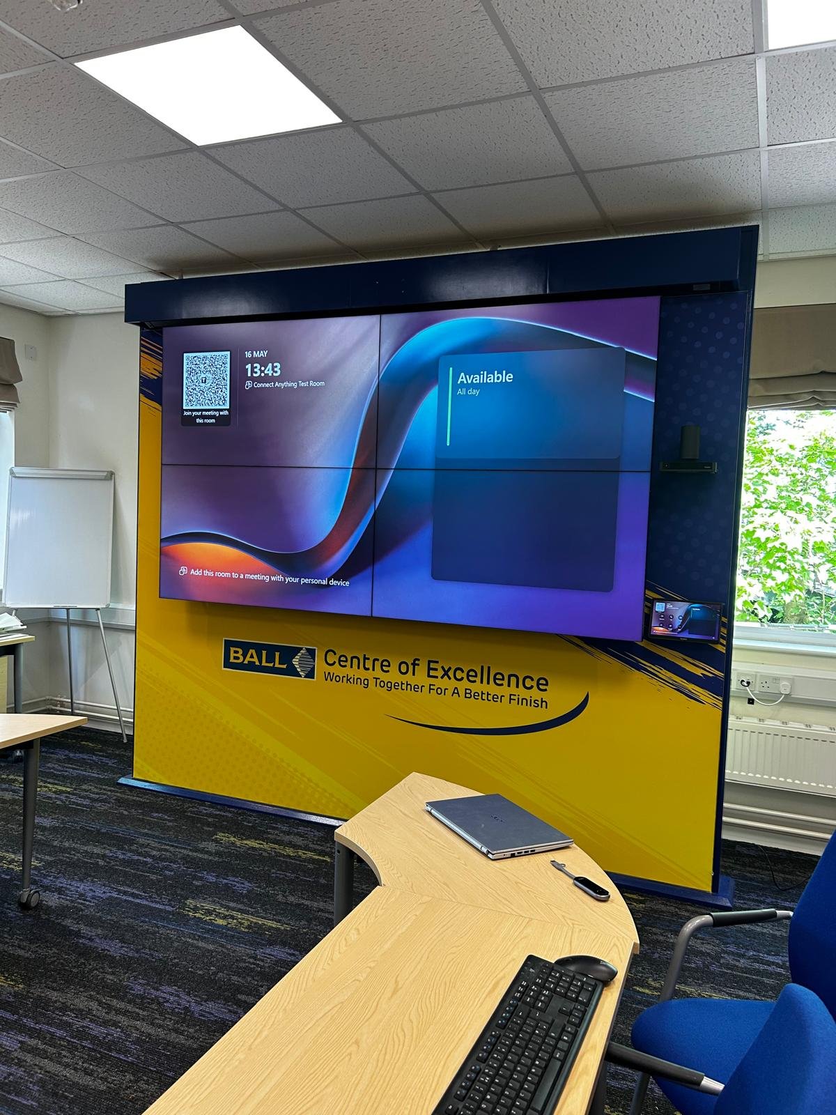 Large digital screen displaying meeting room information at the Ball Centre of Excellence, with a yellow and black sign below it. The room appears to be inside an office or conference room with tables, chairs, and nearby window with greenery outside.