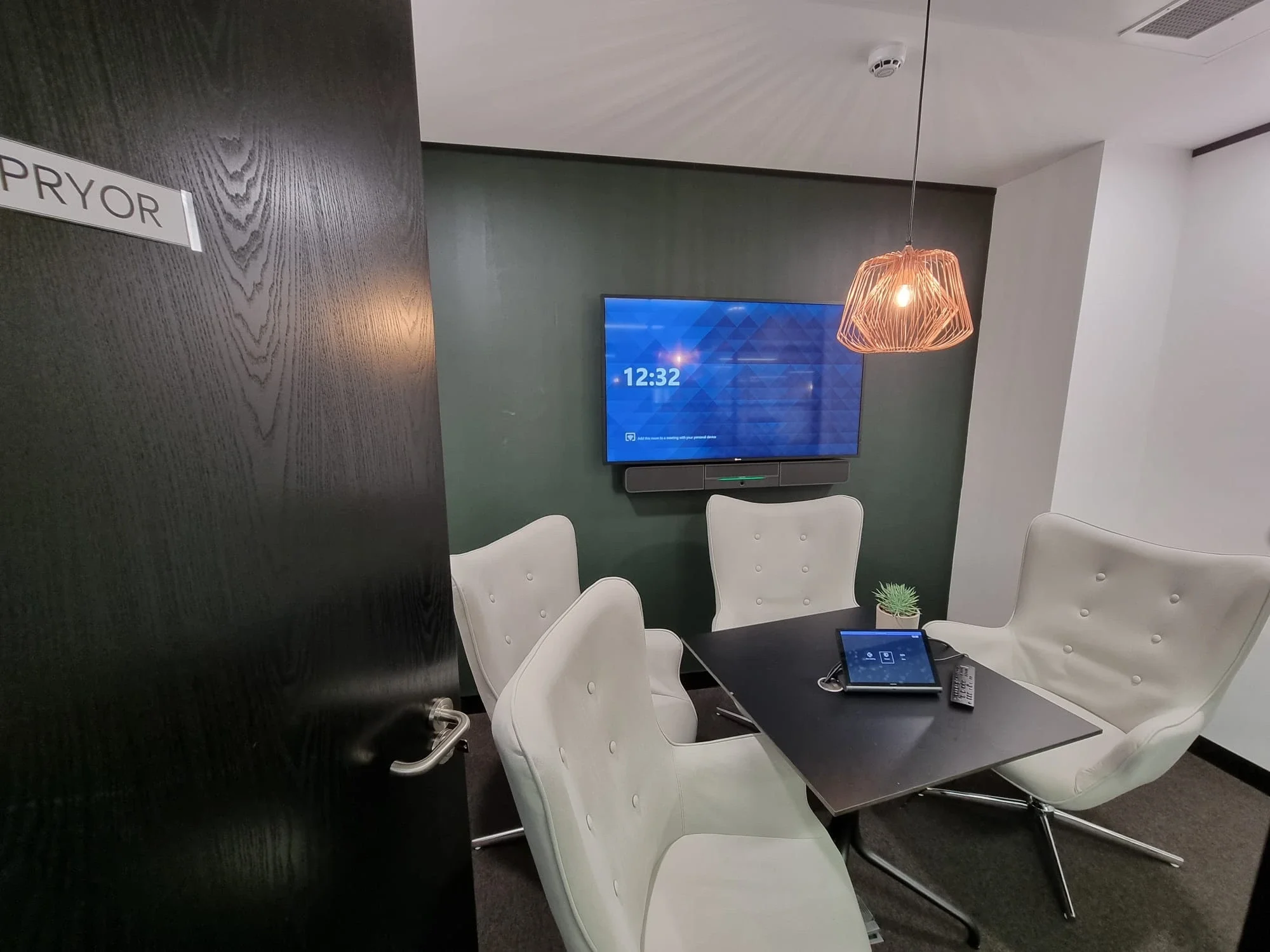 A conference room with a black table surrounded by white chairs, a wall-mounted digital clock showing 12:32, a large flat-screen TV displaying a blue pattern, a light fixture with a wireframe design, a small potted plant, and a tablet with a pen and remote on the table. There is a black door on the left with a sign that reads 'PRYOR' and a ceiling vent.
