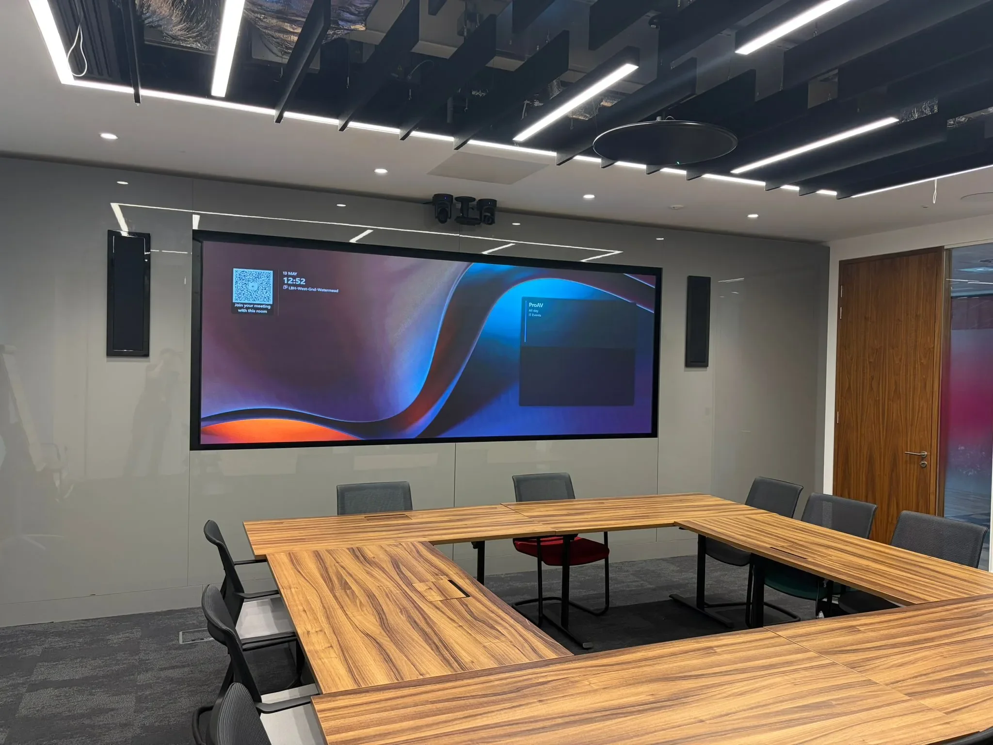 A modern conference room with a large wall-mounted digital display showing meeting information, a wooden conference table with chairs, and ceiling-mounted lighting and ventilation fixtures.