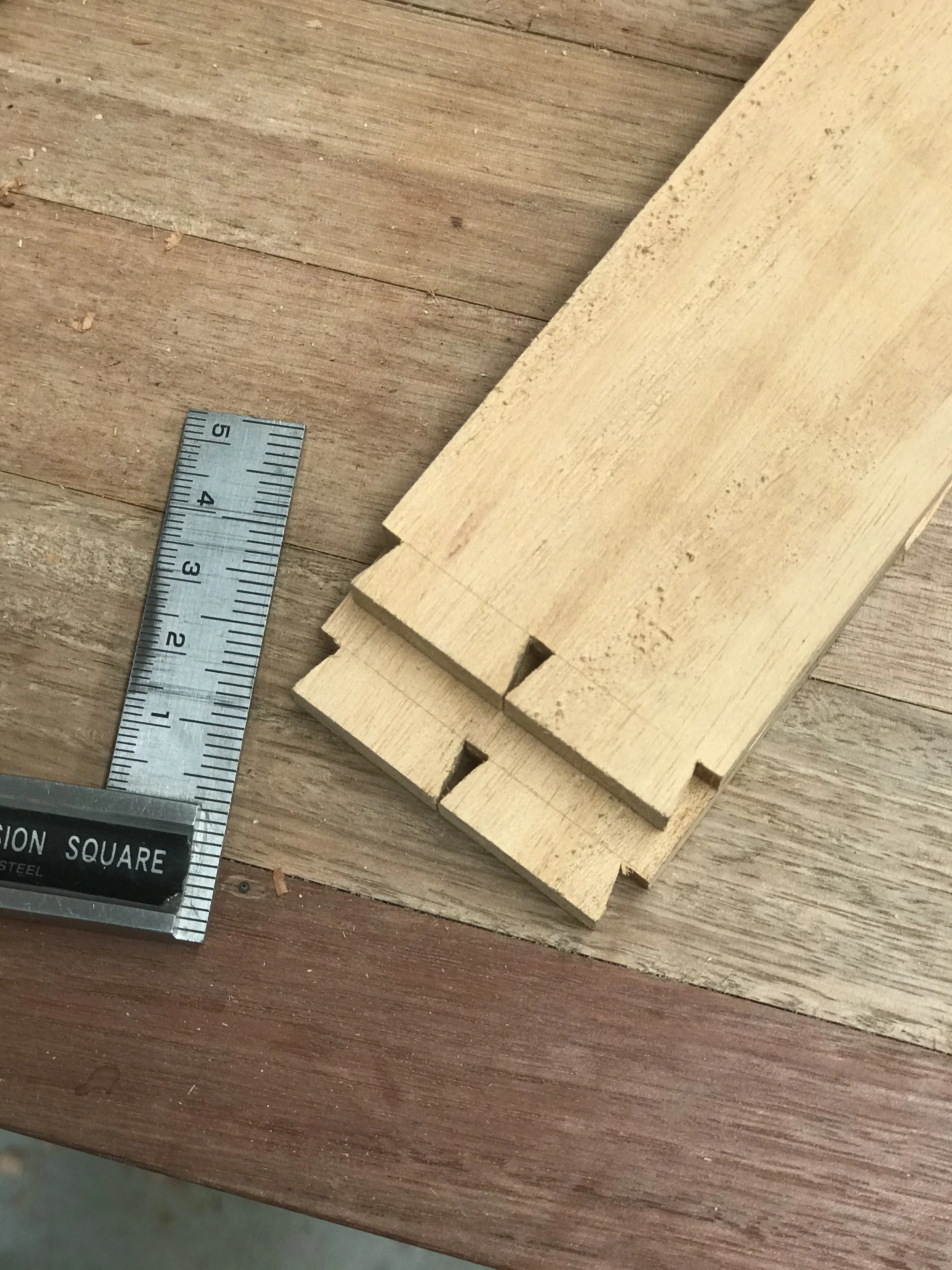 Close-up photo of several light-colored wooden pieces with a square pattern on the ends, placed on a wooden floor. A metal ruler with centimeter markings is partially visible near the corner of the image.