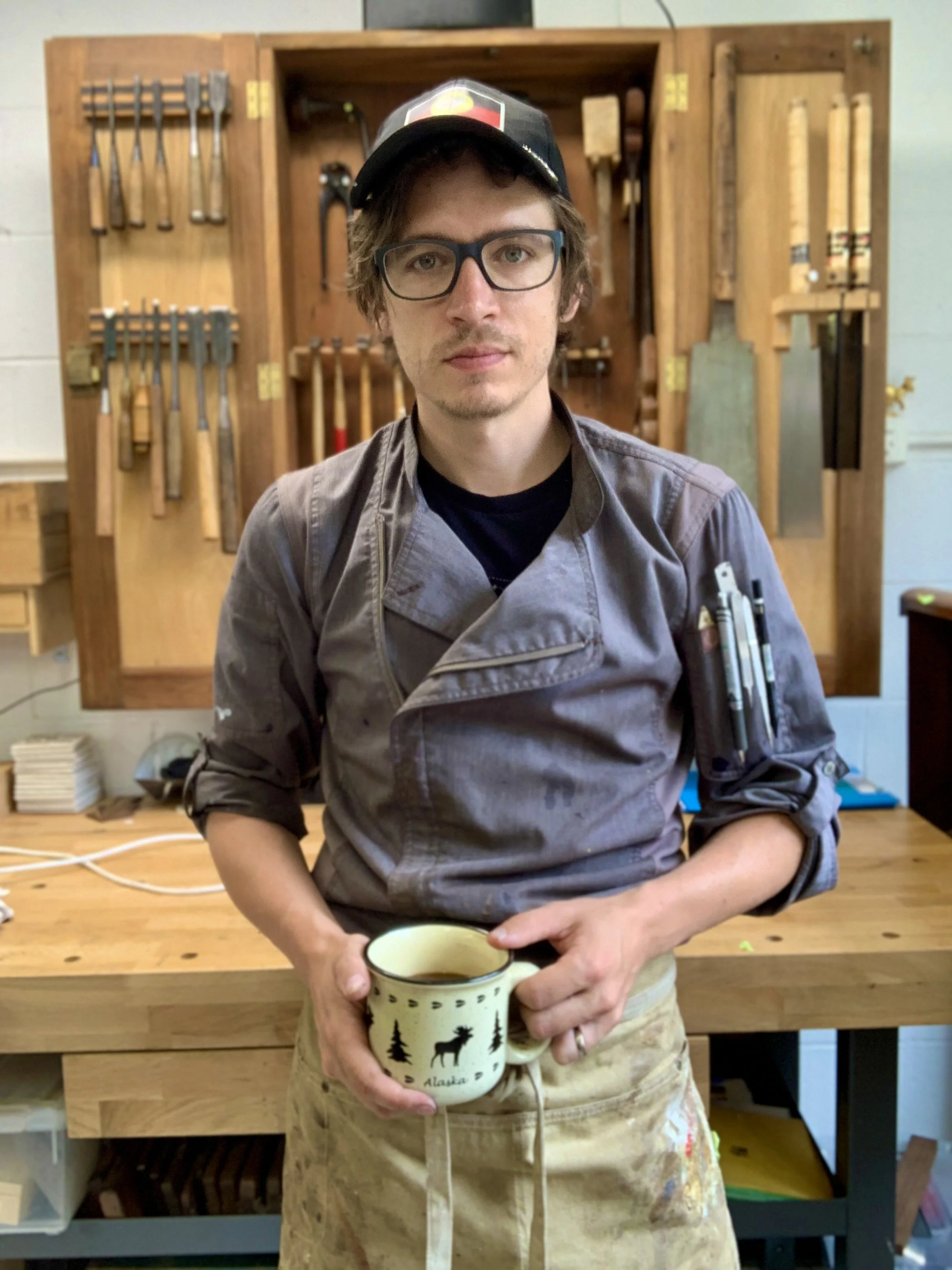 A man wearing glasses, a black cap, and a gray apron holding a mug with a moose and trees design, standing in a woodworking studio with tools on a wooden wall behind him.
