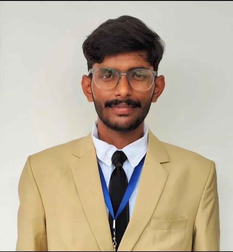 Young man with glasses, dark hair, and a beard wearing a beige blazer, white shirt, a black tie, and a blue lanyard against a plain white background.