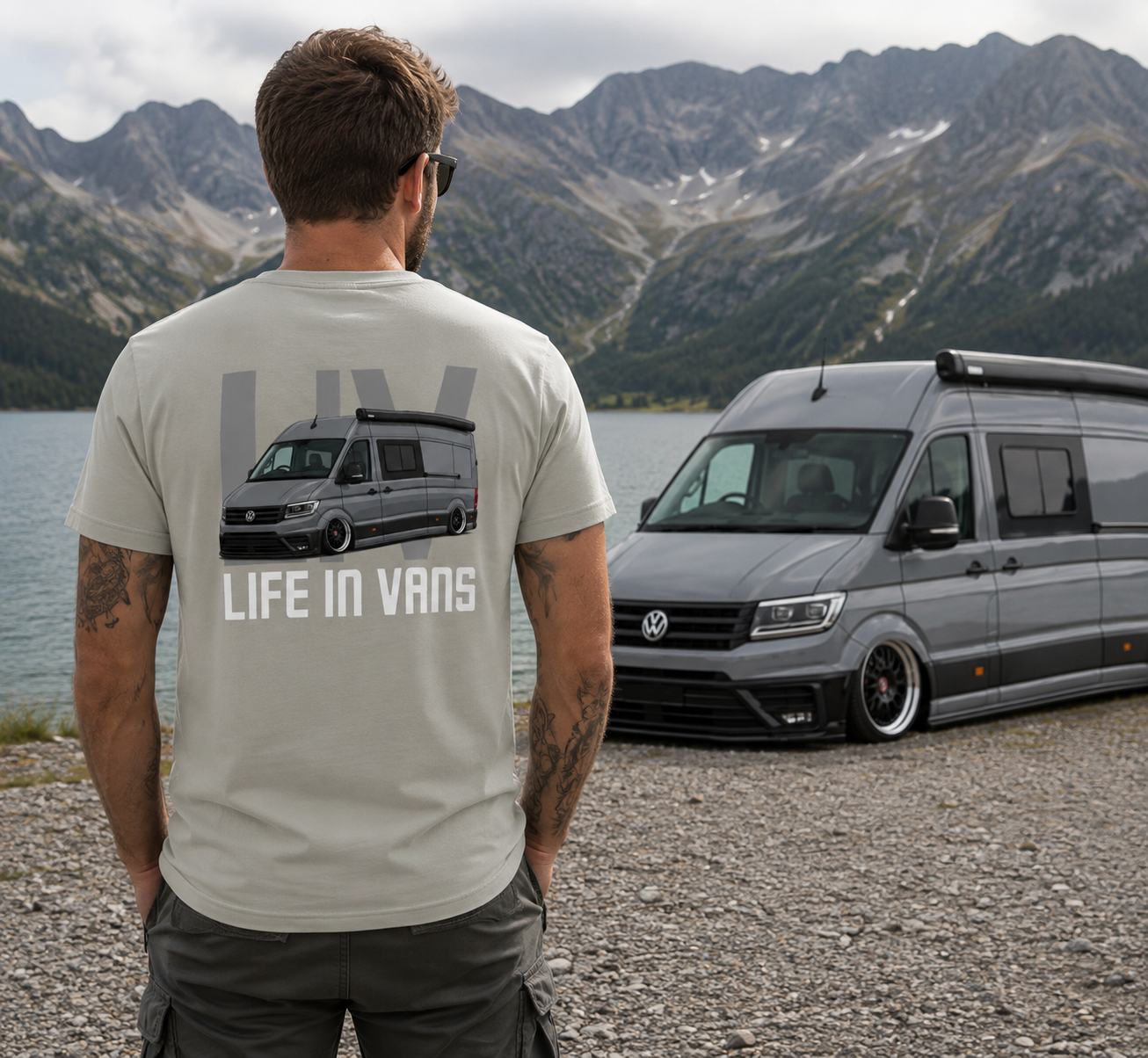 A man wearing sunglasses and a beige T-shirt with a graphic of a camper van and the words "LIFE IN VANS" stands by a lake with mountains in the background. There are two gray camper vans parked nearby.