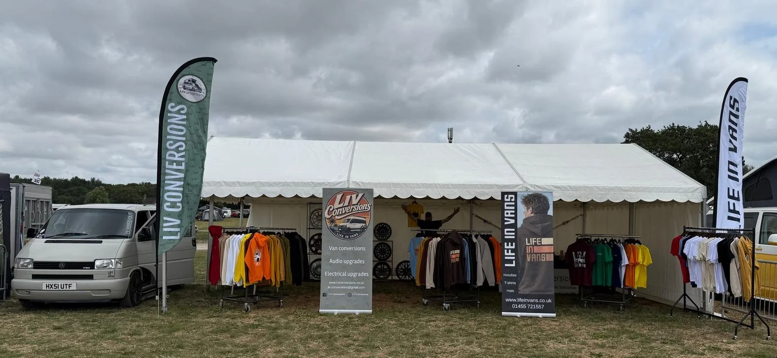 A trade show booth with clothing racks displaying hoodies, t-shirts, and hats under a white tent. Banners read 'LIFE IN VANS' and 'LIVE CONVERSIONS' and a sign promotes van conversions, audio upgrades, and electrical upgrades. There is a gray van parked to the left and stormy clouds overhead.
