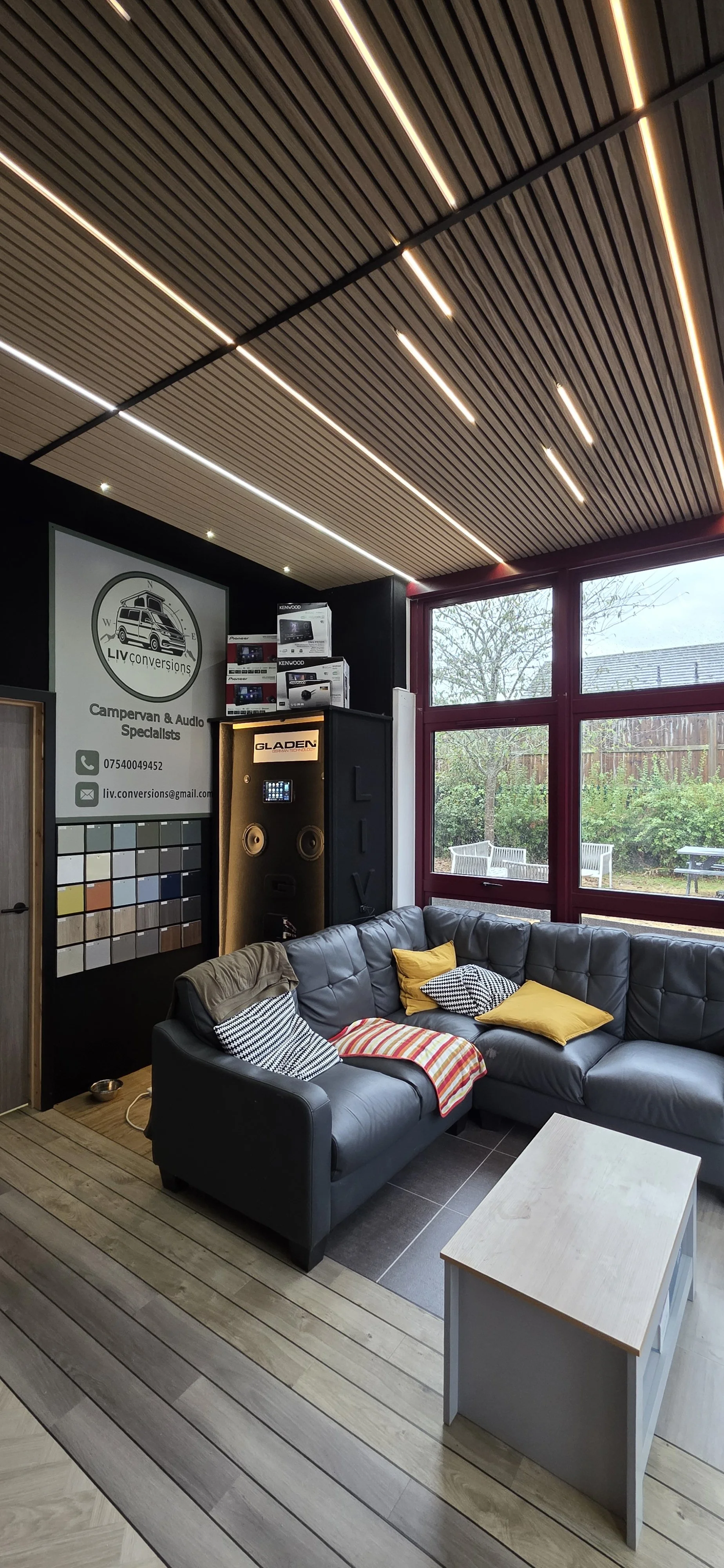 Interior of a modern lounge area with a black leather sectional sofa with colorful cushions, a small white table, large windows showing an outdoor garden, a wall with a sign for LIV conversions, and several audio equipment boxes on shelves.