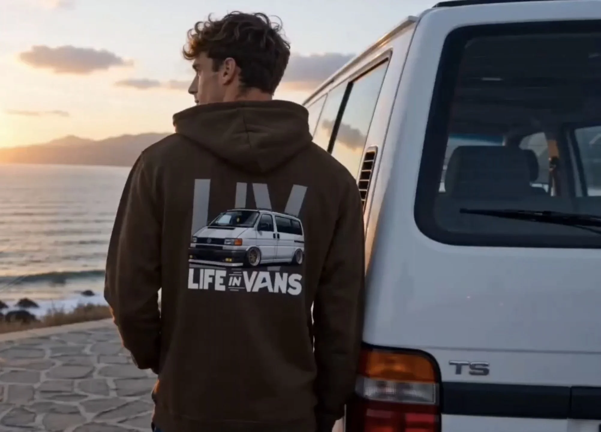 Young man with curly hair looking away, standing next to a white van on a paved waterfront road during sunset. Wears a brown hoodie with a graphic of a van and the text 'LIFE IN VANS' on the back.