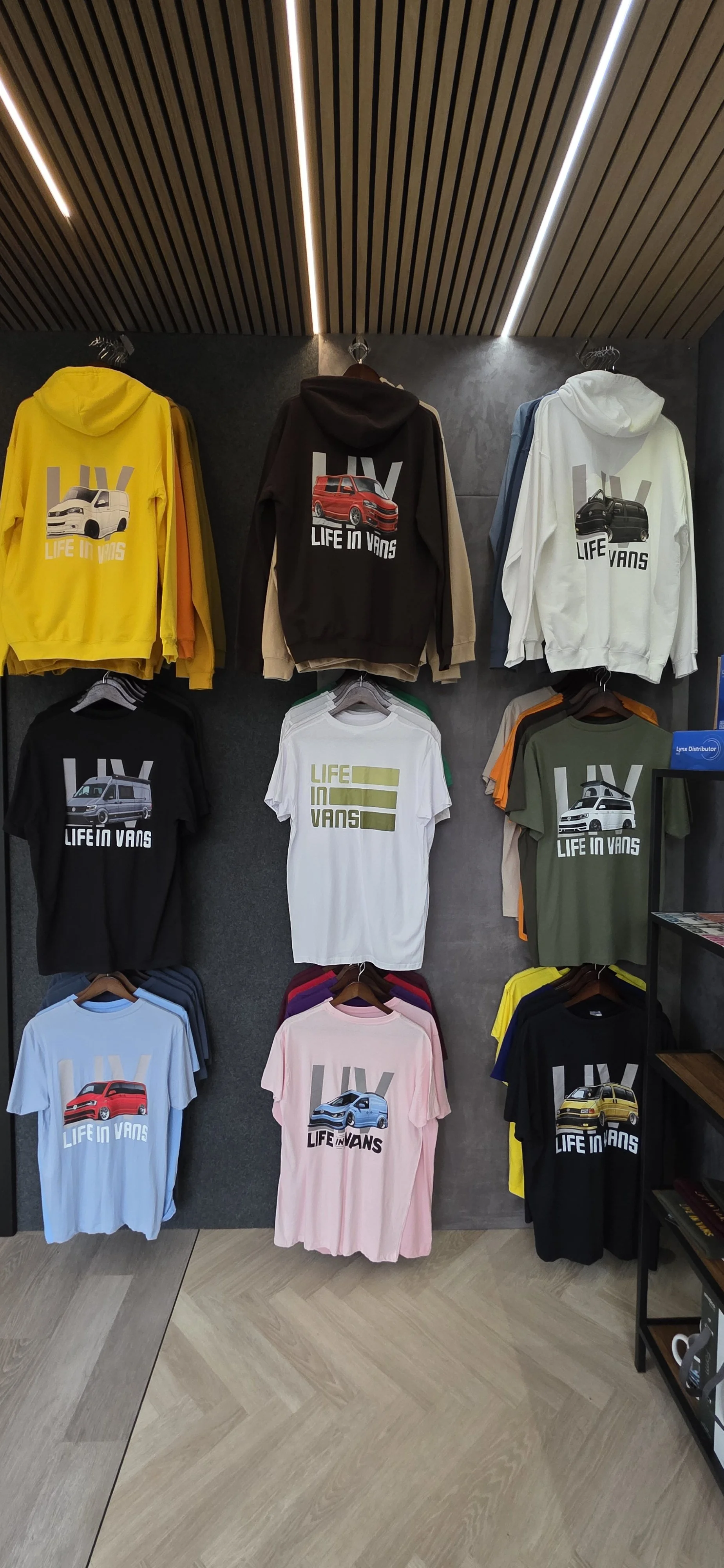 Display of colorful t-shirts and hoodies with car graphics and the slogan 'LIFE IN VANS' on a store wall.