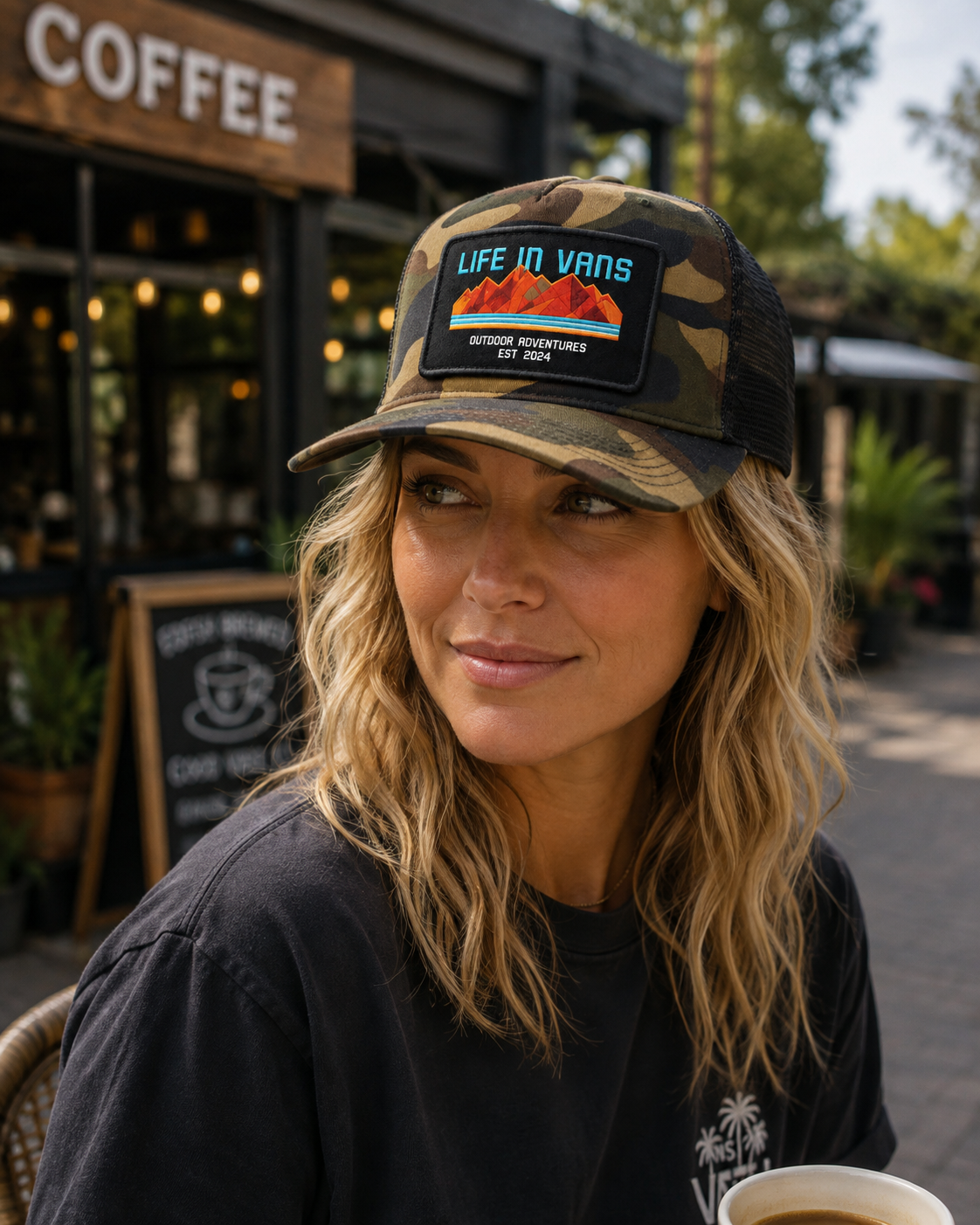 A woman with blonde curly hair wearing a camouflage baseball cap with a 'Life in Vans' patch and a black t-shirt, sitting outdoors at a cafe.