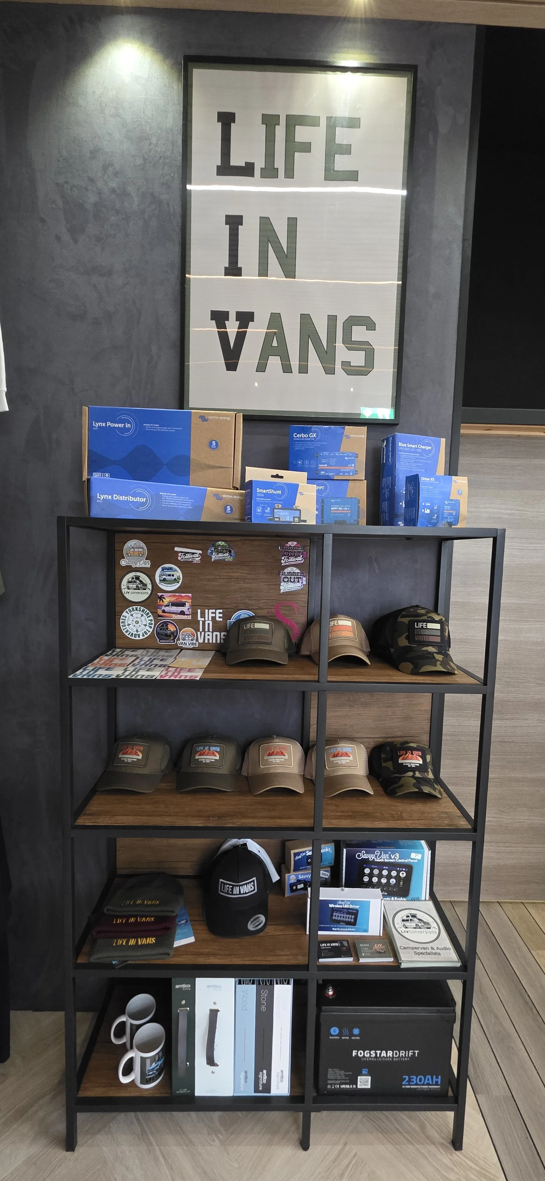 Shelving unit with automotive and lifestyle merchandise including hats, mugs, batteries, gadgets, and accessories, with a sign reading "Life in Vans" and a framed sign at the top that says "LIFE IN VANS."
