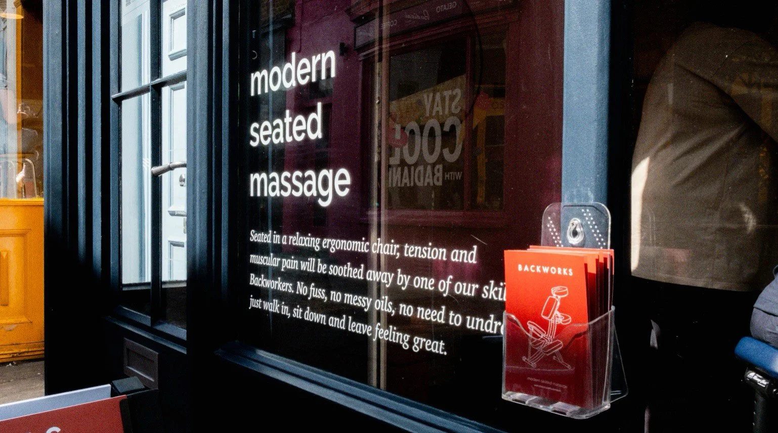 A storefront window advertising modern seated massage with white text on a black background, describing massage benefits, and a red brochure stand labeled "Backworks" with a diagram of a massage chair inside.
