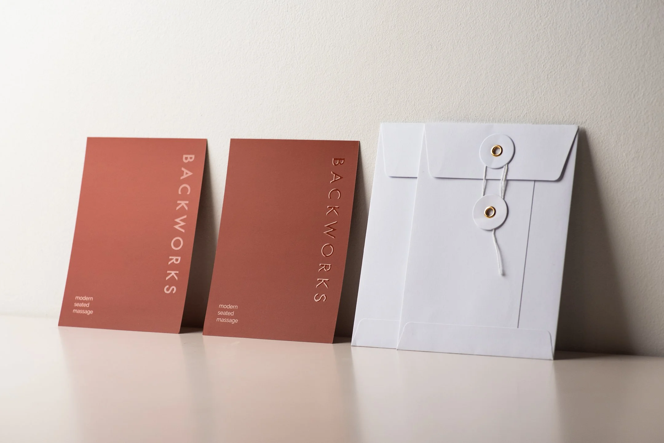 Two reddish-brown cards with white text reading 'BACKWORKS' stand upright on a white surface, with a white envelope with a string seal beside them.