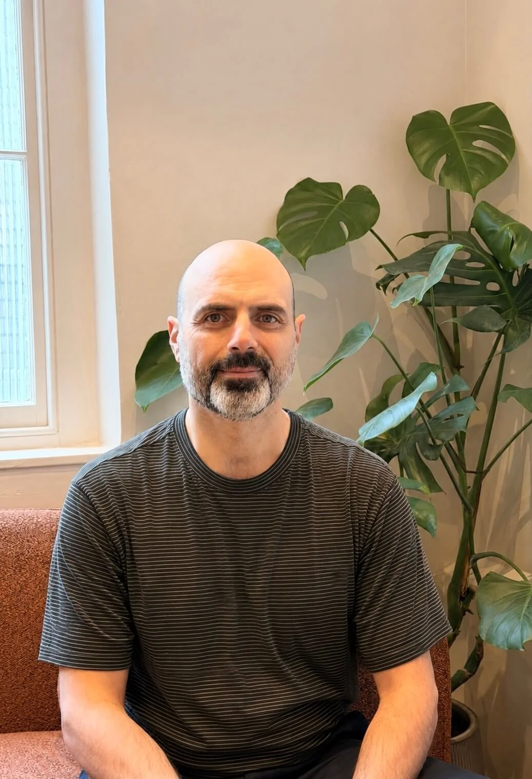 A man with a beard and bald head sitting on a brown textured couch in front of a beige wall with large leafy green plants beside him, near a window with frosted glass.