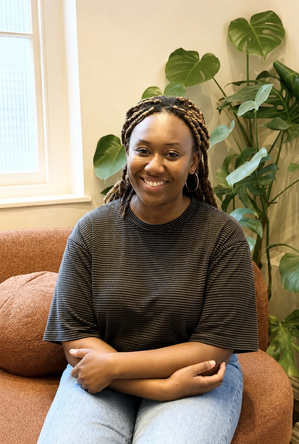 A woman with braided hair, wearing hoop earrings and a black striped T-shirt, sitting on a rust-colored couch with arms crossed, smiling, in a room with a window and green leafy plants in the background.