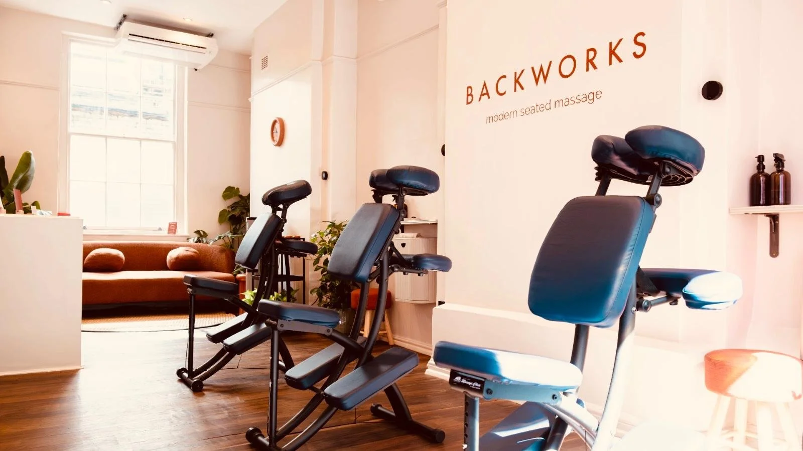 Interior of a massage studio with blue massage chairs lined up, a wooden bench, and a  large glass window. The walls are painted light, with a sign that reads 'BACKWORKS modern seated massage,' and there is sunlight coming through the window.