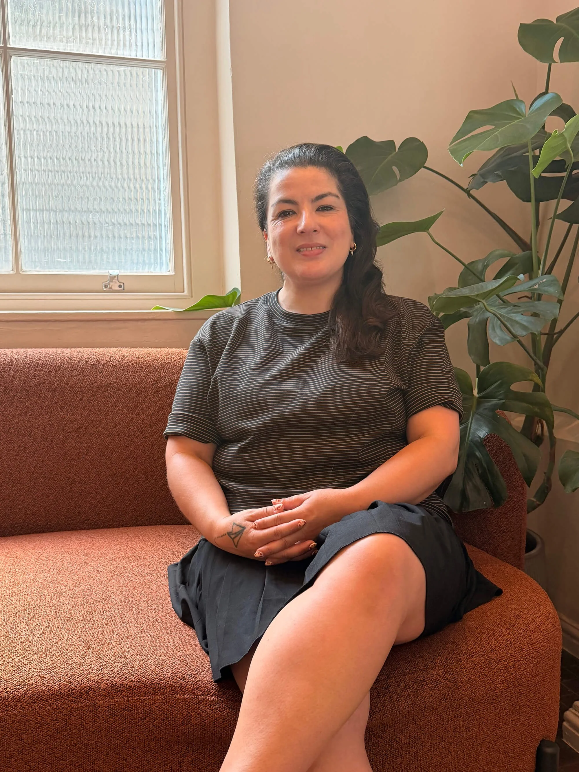 A woman sitting on a reddish-brown couch near a large window with textured glass, next to a green leafy plant in a beige wall background. She is wearing a striped shirt and black shorts, with visible tattoos on her left forearm.